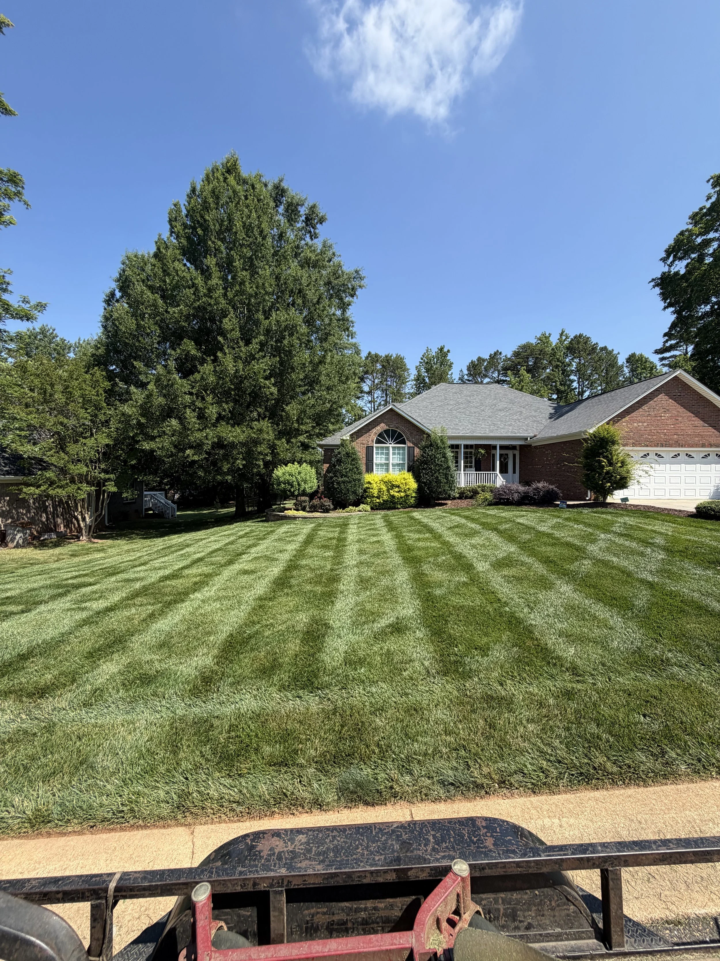 View from a lawn mower of a well-maintained front yard with green grass, shrubs, trees, and a brick house with a gray roof and white garage door under a partly cloudy blue sky.