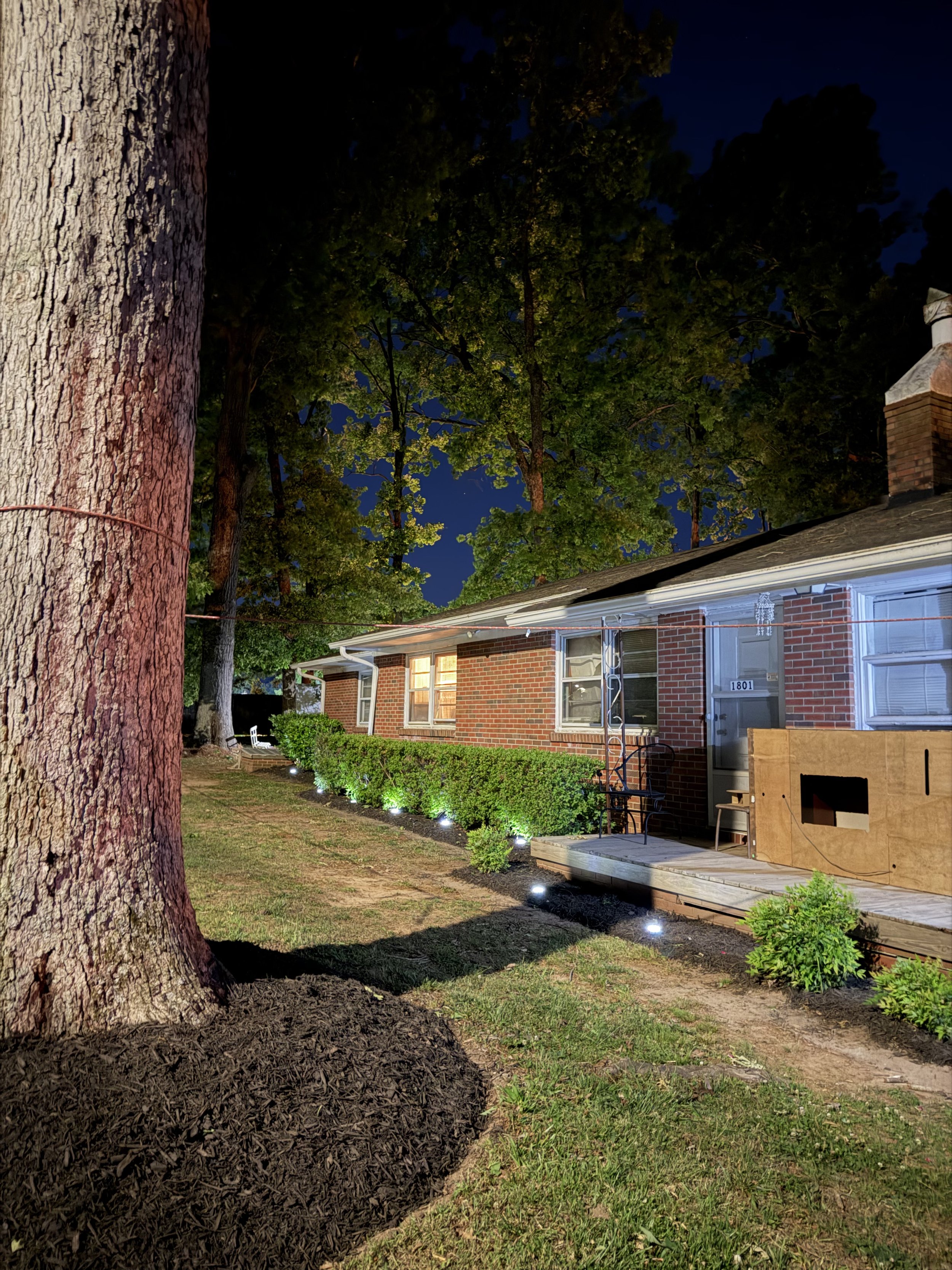 Nighttime view of a backyard with a large tree on the left, a row of small landscape lights along the lawn, and a brick house with several windows illuminated, a porch, and a chimney on the right.