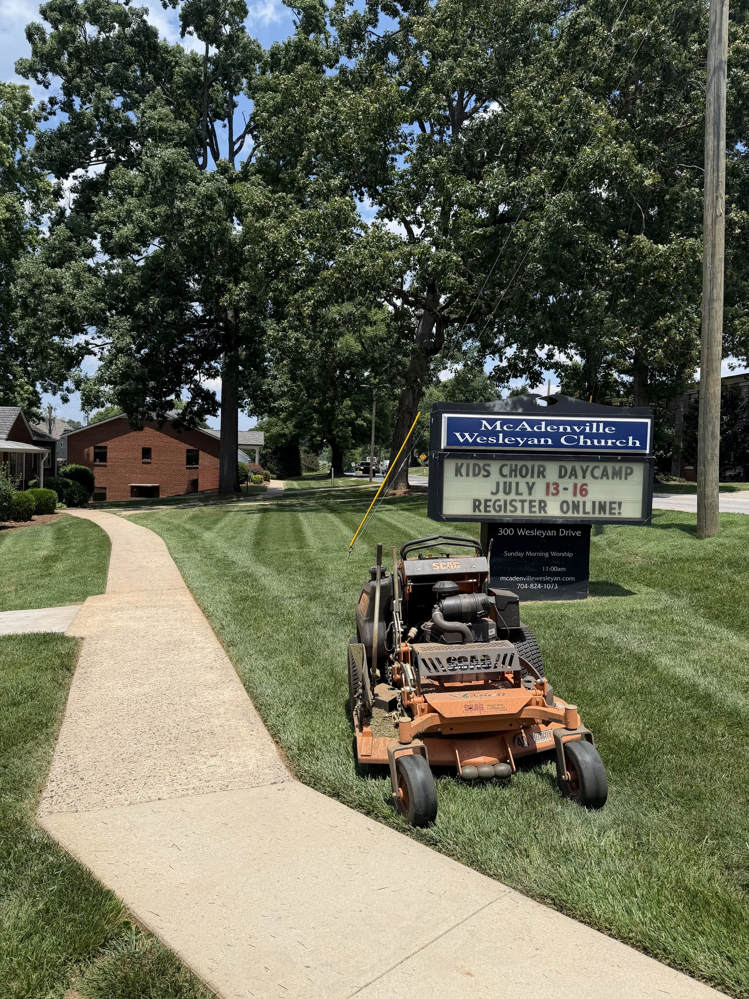 A landscaped sidewalk with a lawn mower in front of a church sign for McAdenville Wesleyan Church, advertising kids choir daycamp from July 13-16, in a suburban area with houses, trees, and a blue sky.