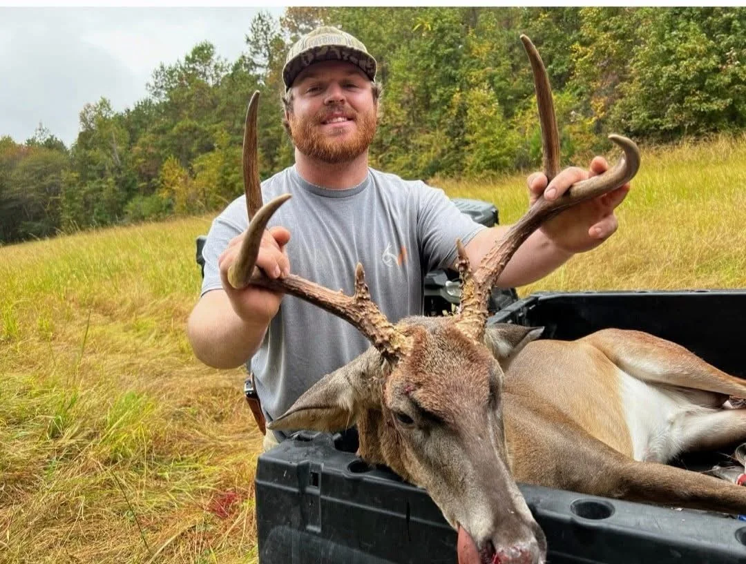 A man with a beard and camouflage cap holding the antlers of a large deer he just hunted, lying in the back of a truck in a field with trees in the background.