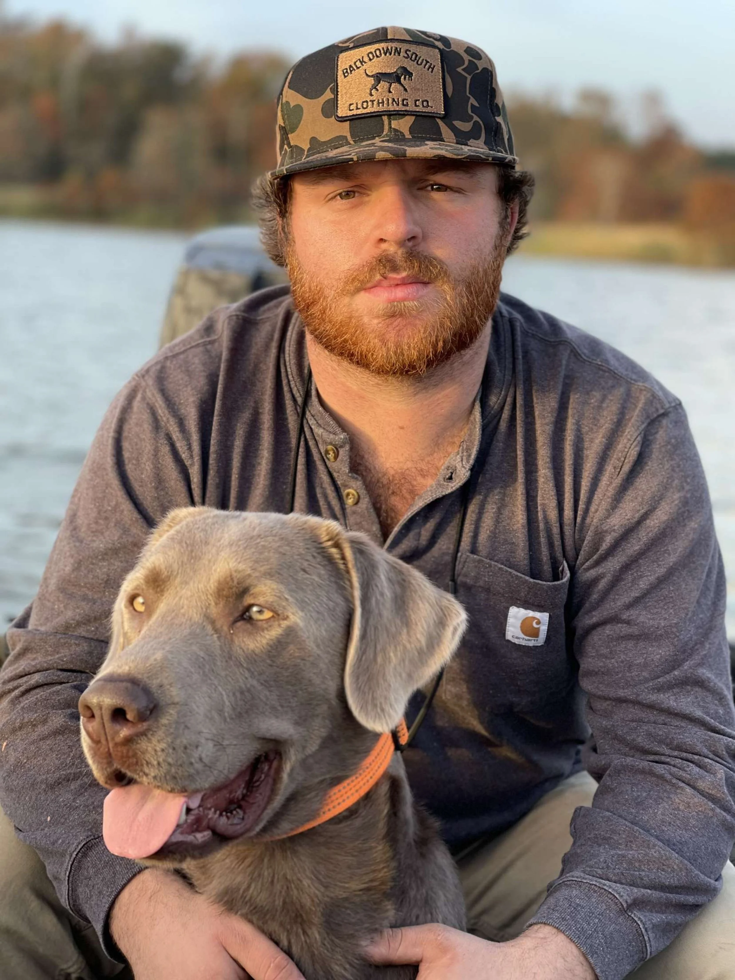 A man with a beard wearing a camouflage baseball cap and a gray Carhartt jacket, holding a happy gray Labrador Retriever puppy outdoors by a body of water with trees in the background