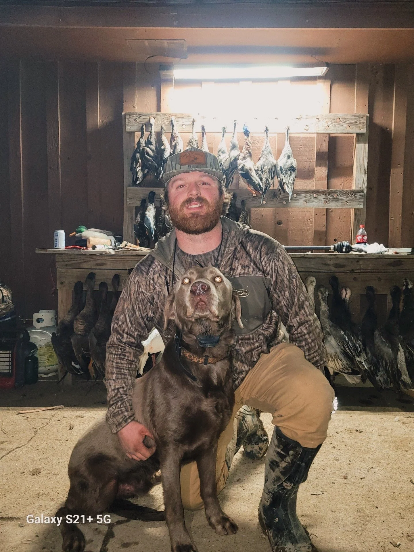 A man in camouflage hunting gear and a cap kneeling next to a large brown dog, inside a wood-paneled room with dead birds hanging on a rack behind them.