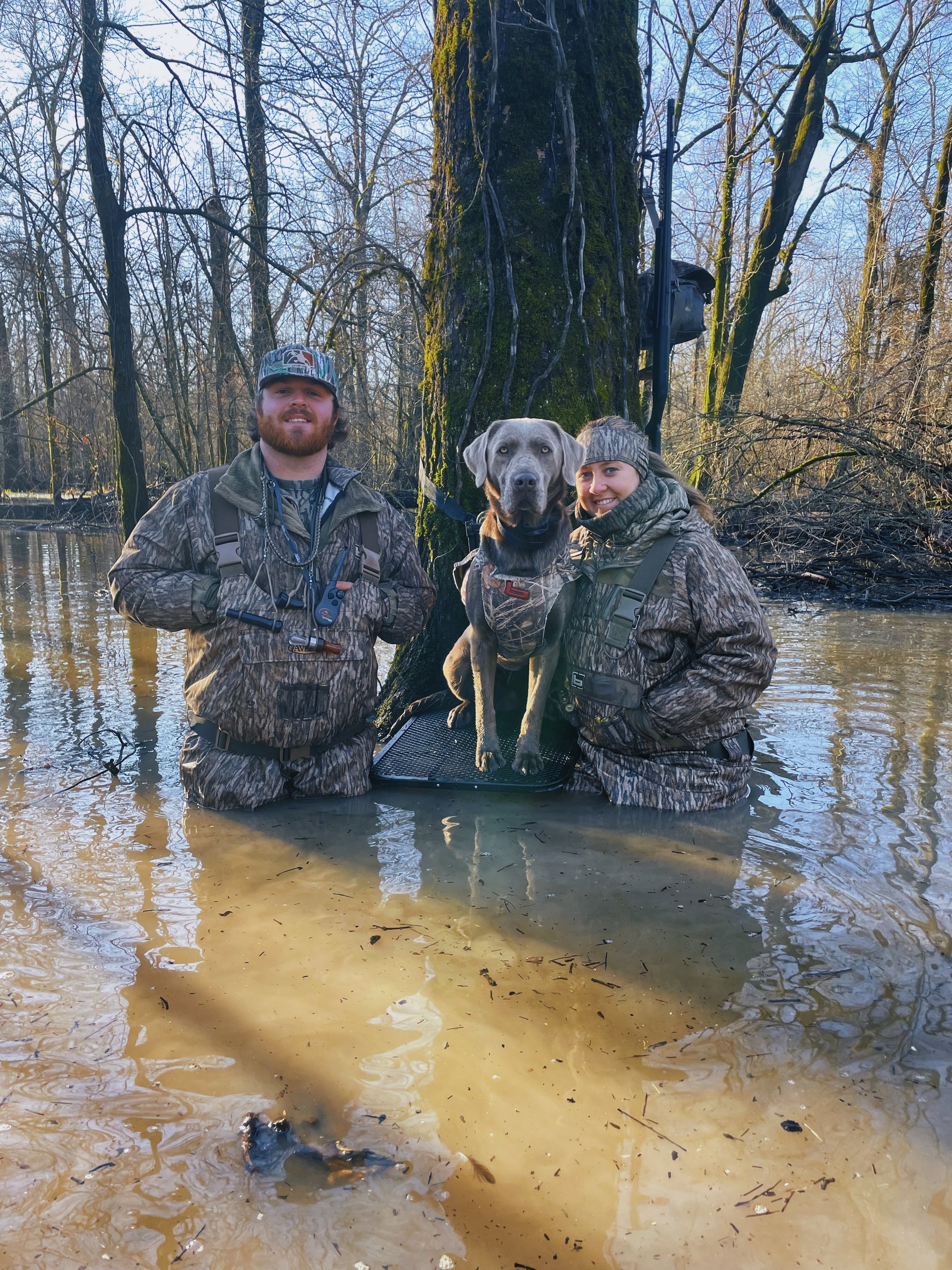 Two people and a dog stand in a flooded area with leafless trees in the background. The man and woman are wearing camouflage clothing and the dog is sitting on a board in the water.