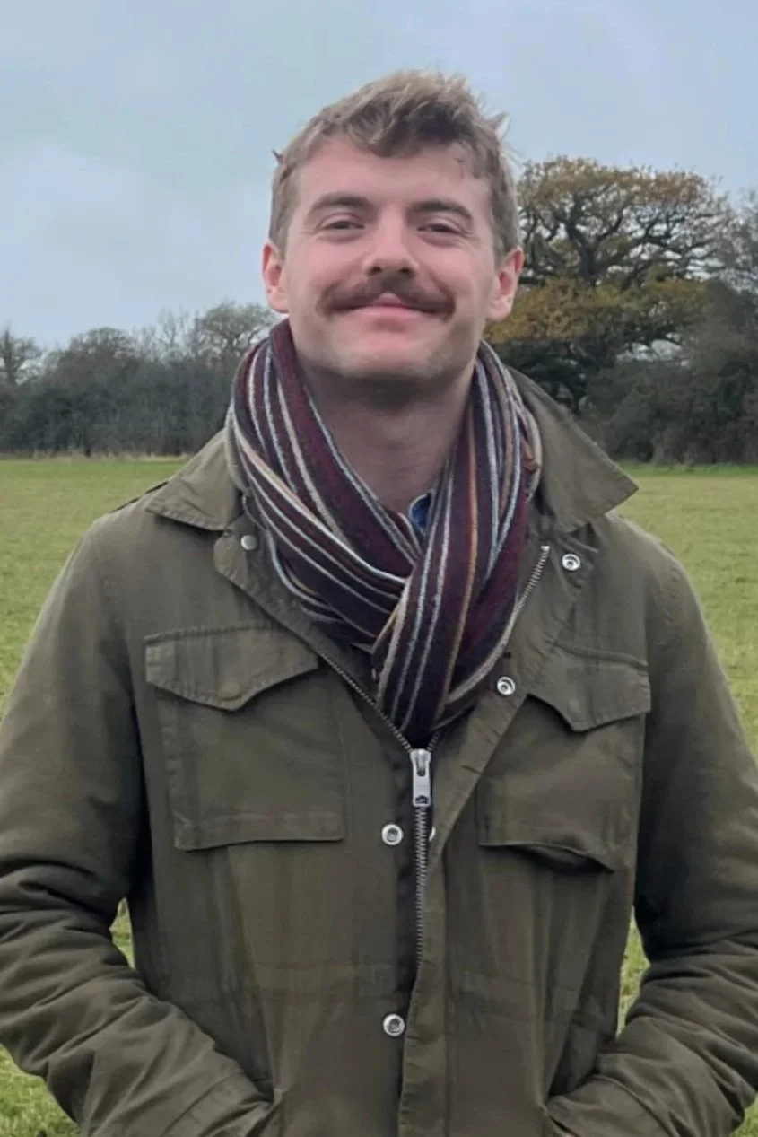 A young man with a mustache smiling outdoors in a field with trees in the background, wearing a green jacket and a striped scarf.