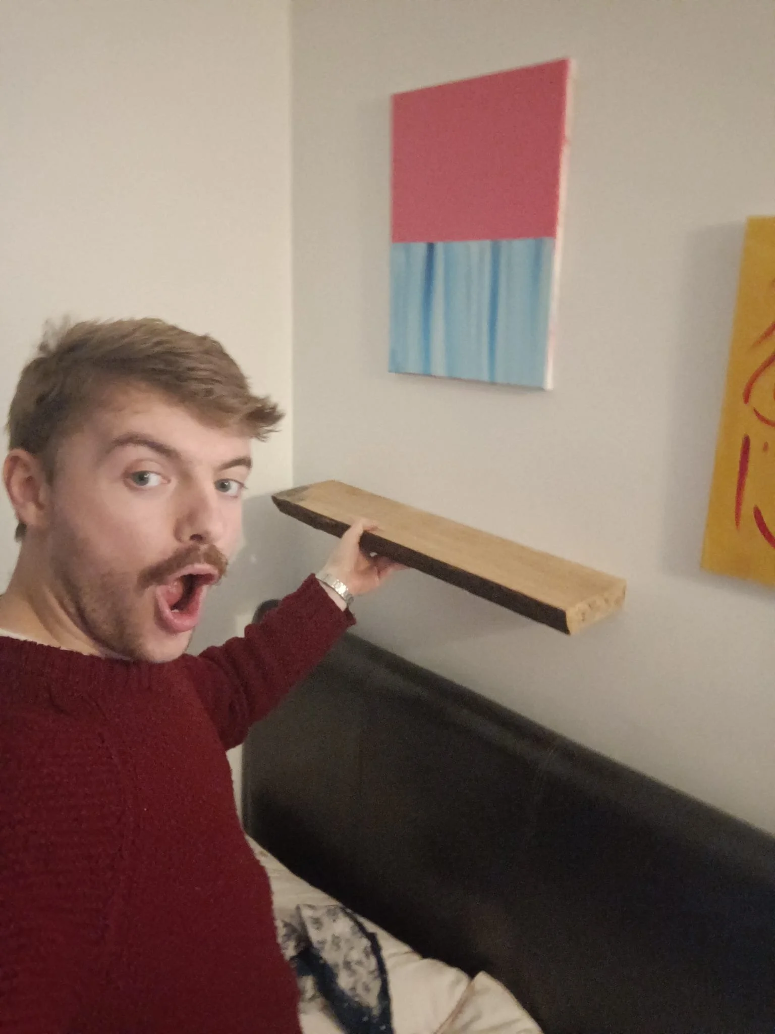 A man with light brown hair and a mustache, wearing a red sweater, holding a wooden plank while standing next to a bed with a black headboard, and appears surprised or excited.