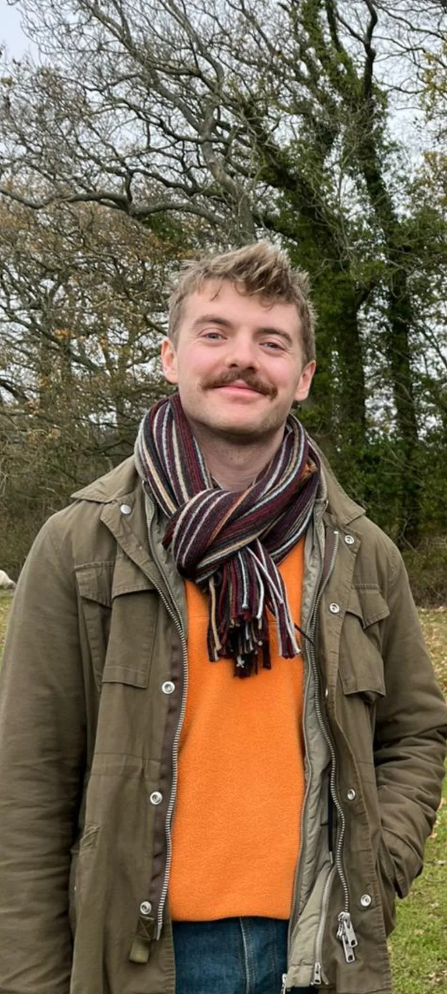 A young man with a mustache and short light brown hair, wearing an orange sweater, green jacket, and a multicolored striped scarf, standing outdoors in front of leafless trees.