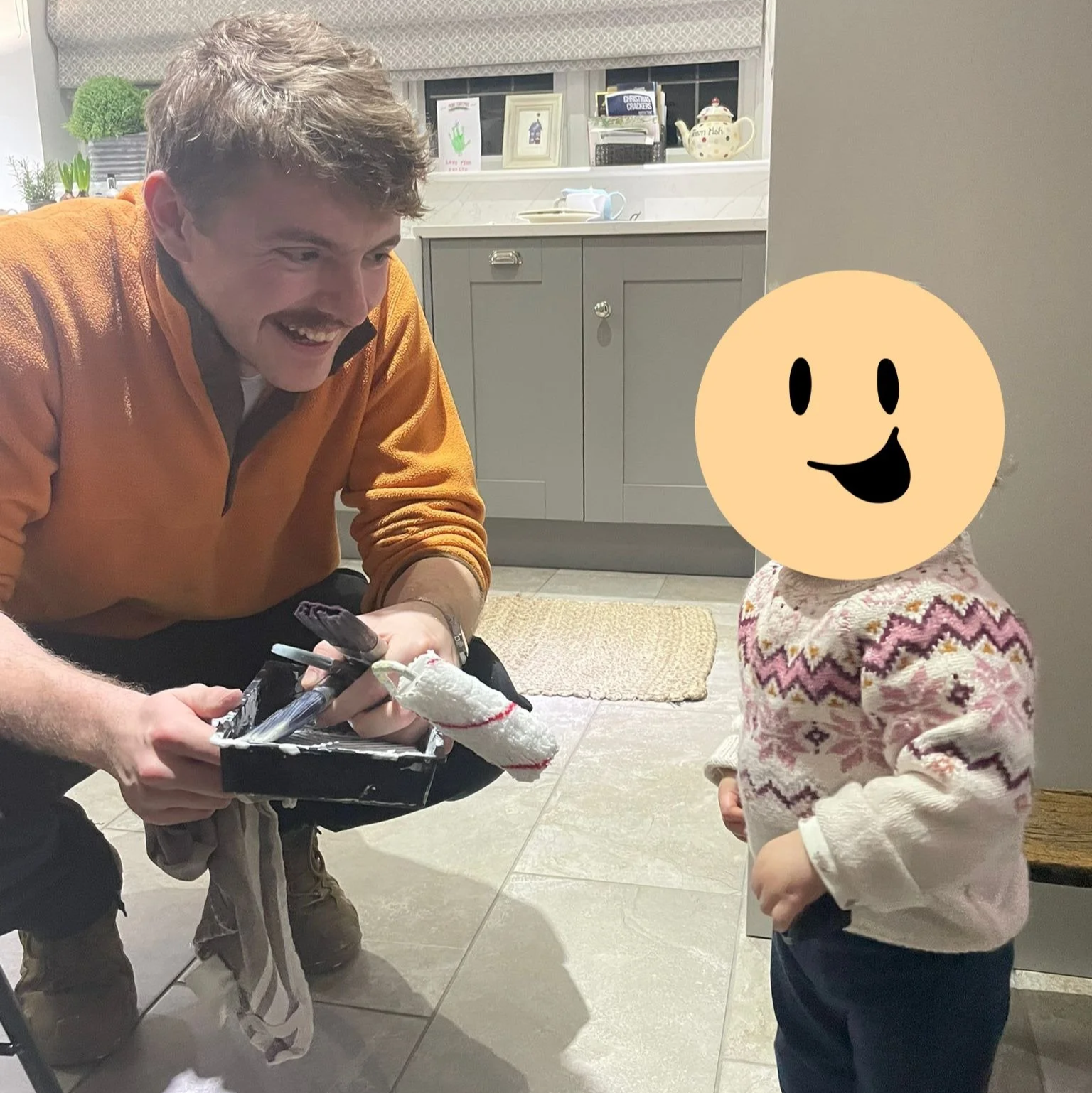 A man kneeling and smiling, holding a tray, while a young girl with a smiley face mask on her face stands nearby in a kitchen.