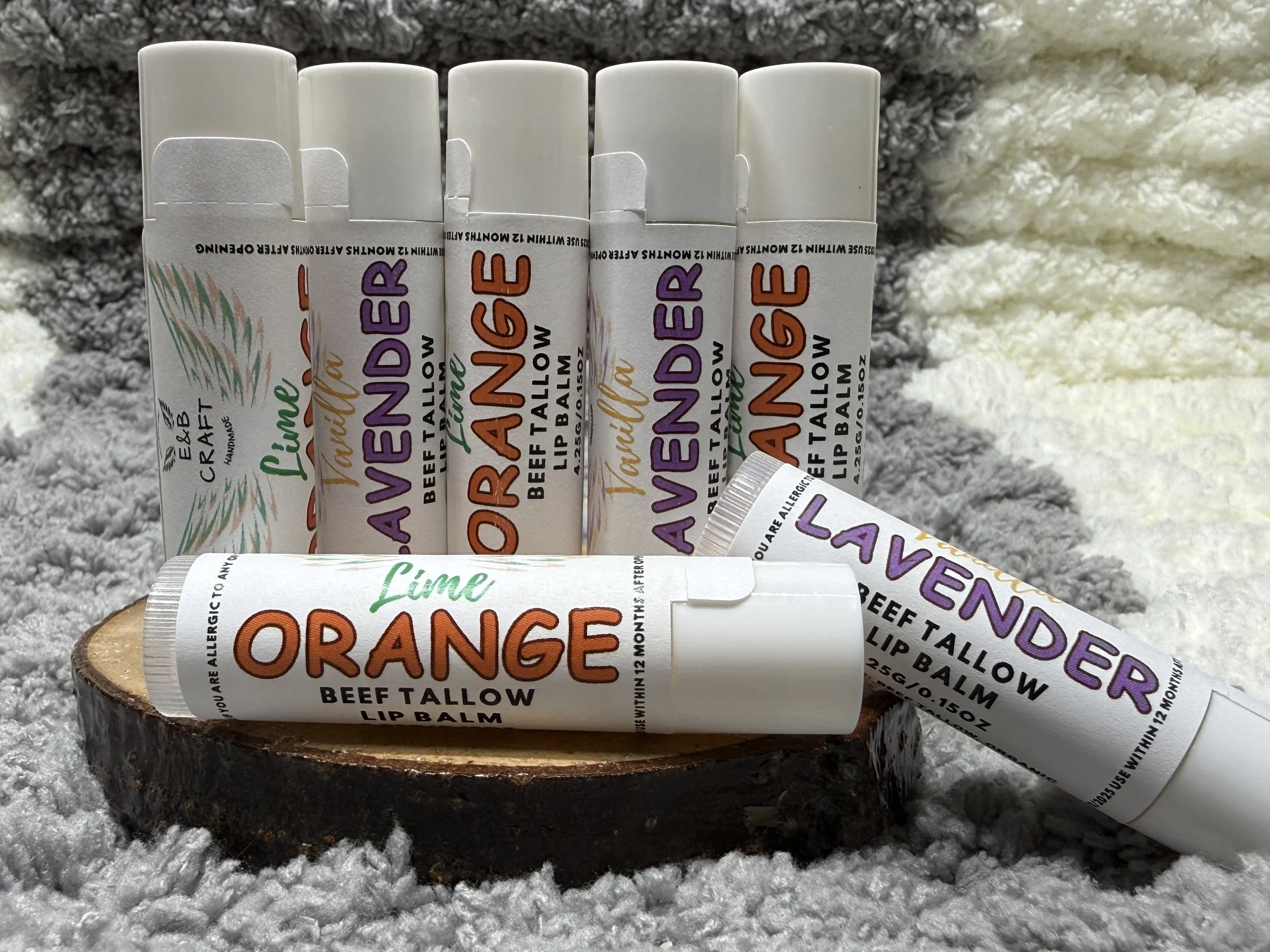 A collection of lip balm tubes with flavors lavender, lime, and orange arranged on a wooden surface with a fluffy gray and white background.