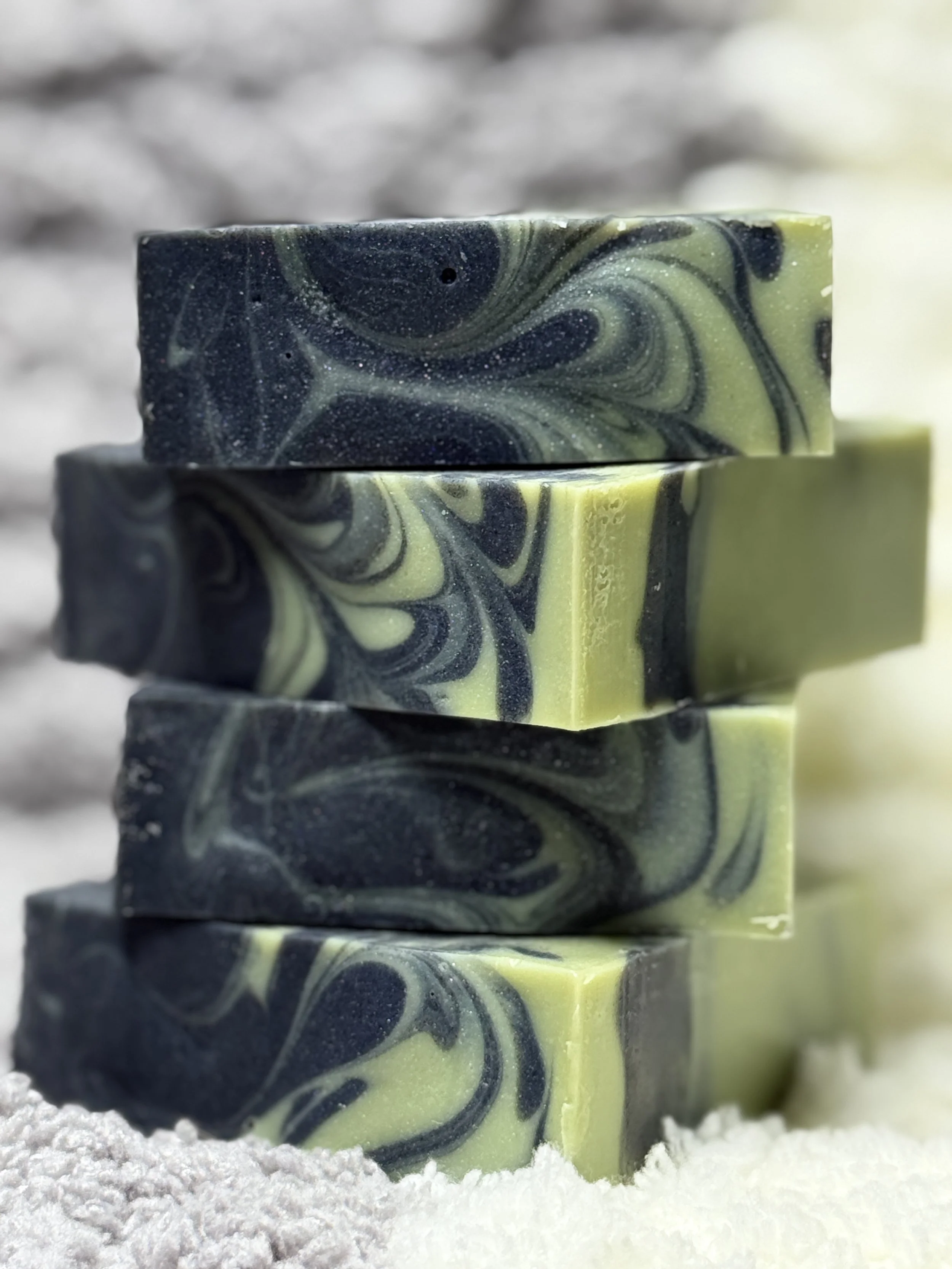 Stack of four swirl-colored soap bars, black and green with marbled patterns, on a snowy surface with a blurred gray background.