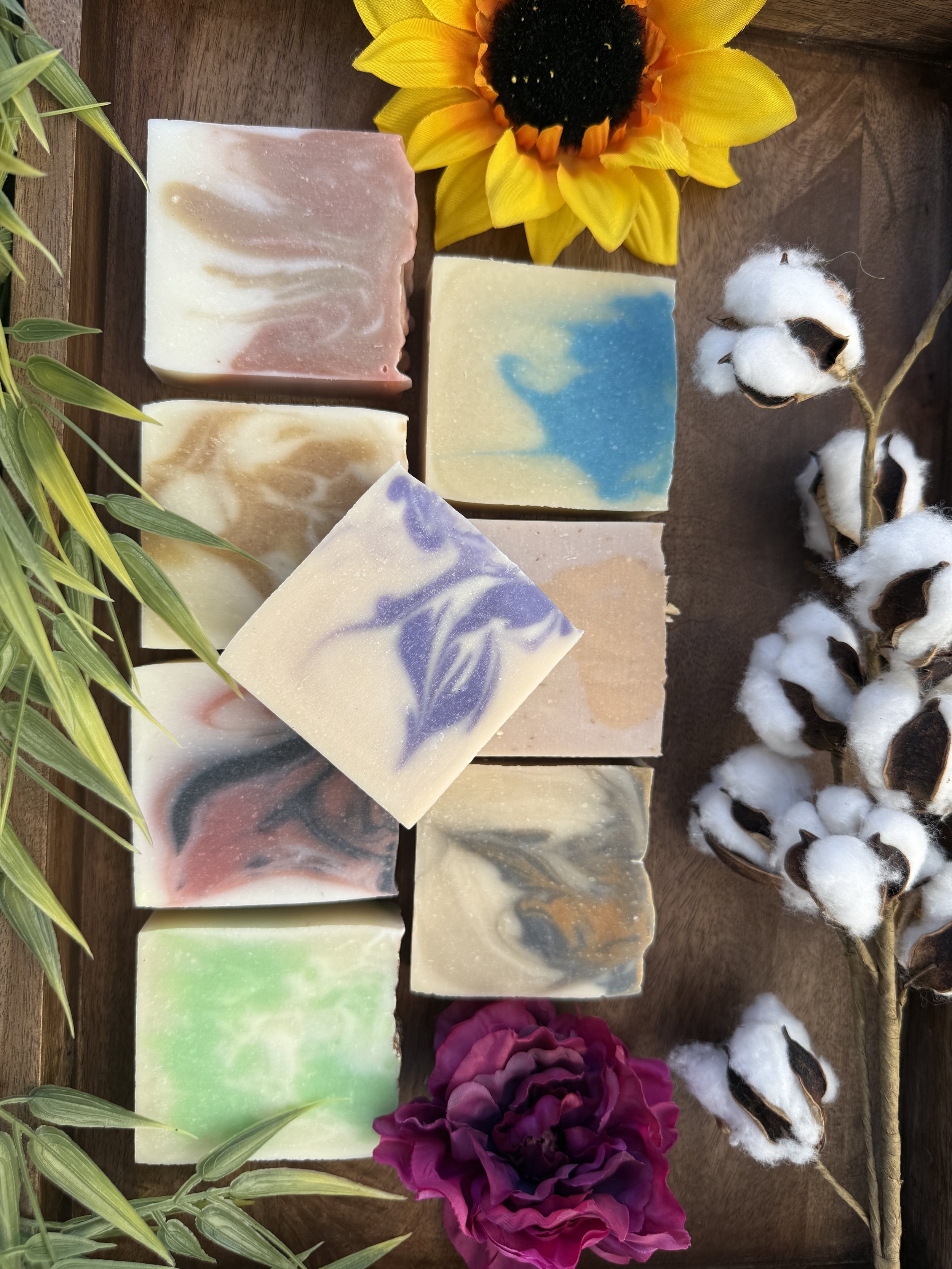 Assorted handmade soaps with colorful marbled patterns on a wooden surface, surrounded by flowers and cotton branches.