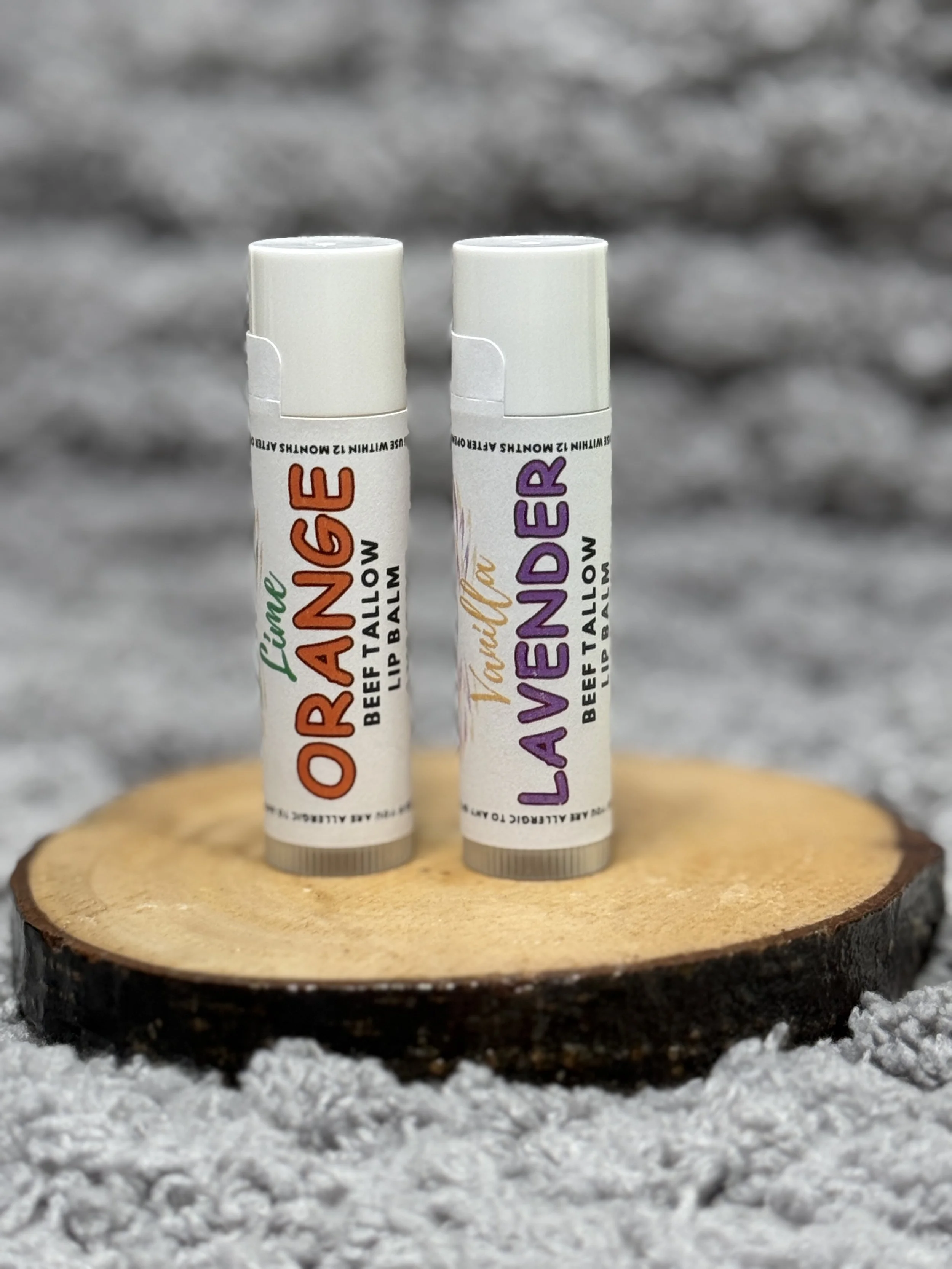 Two lip balm tubes, one labeled 'Orange' in orange and green text and the other labeled 'Lavender' in purple and gold text, stand upright on a wooden circular surface against a blurred gray background.