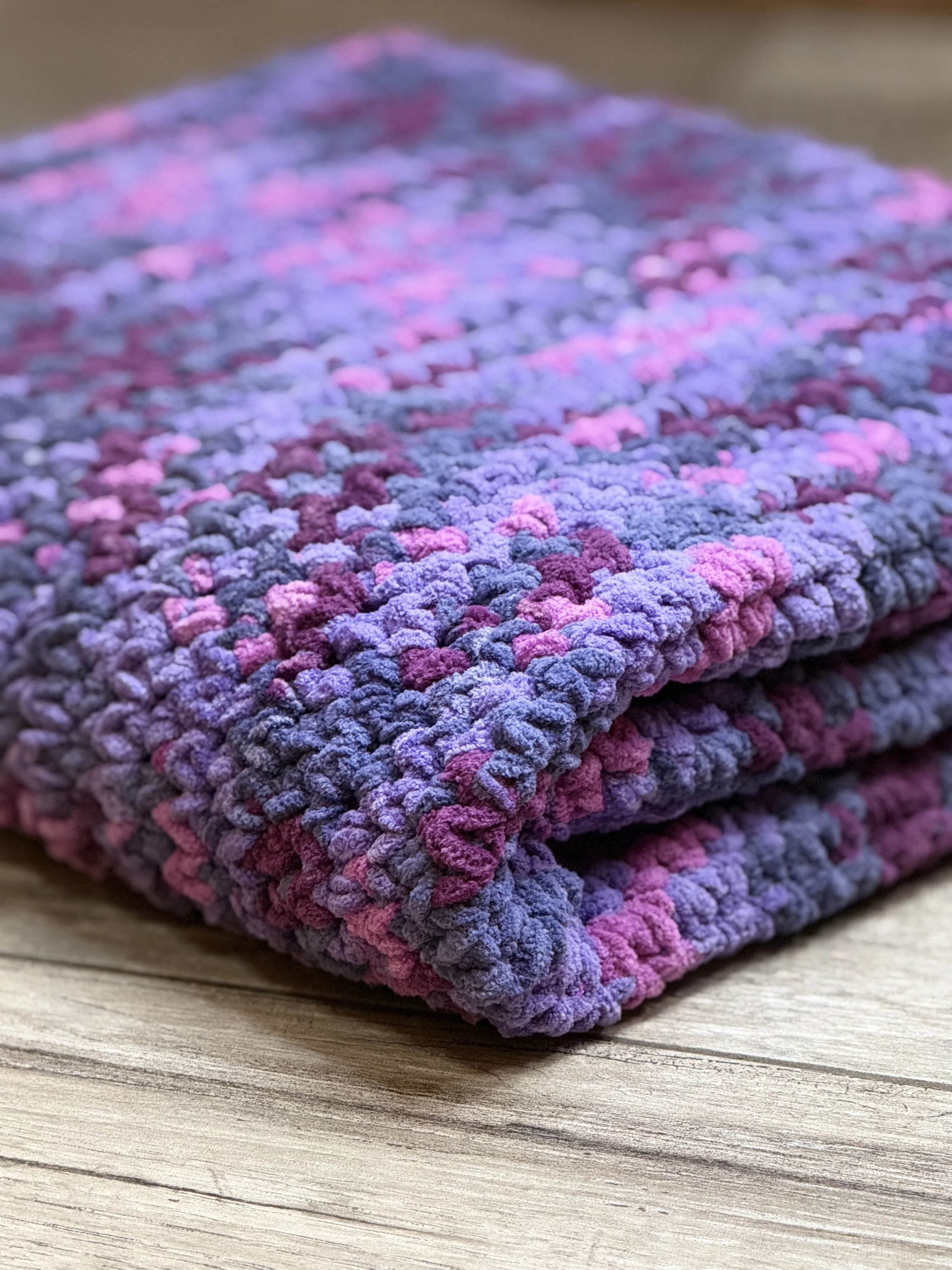 Close-up of a folded multicolored chenille blanket with purple and pink hues on a wooden surface.