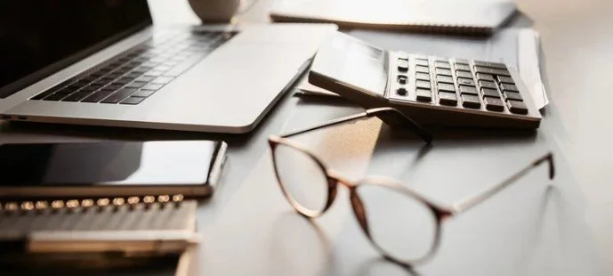 A workspace desk with a laptop, smartphone, calculator, notebook, and a pair of eyeglasses.