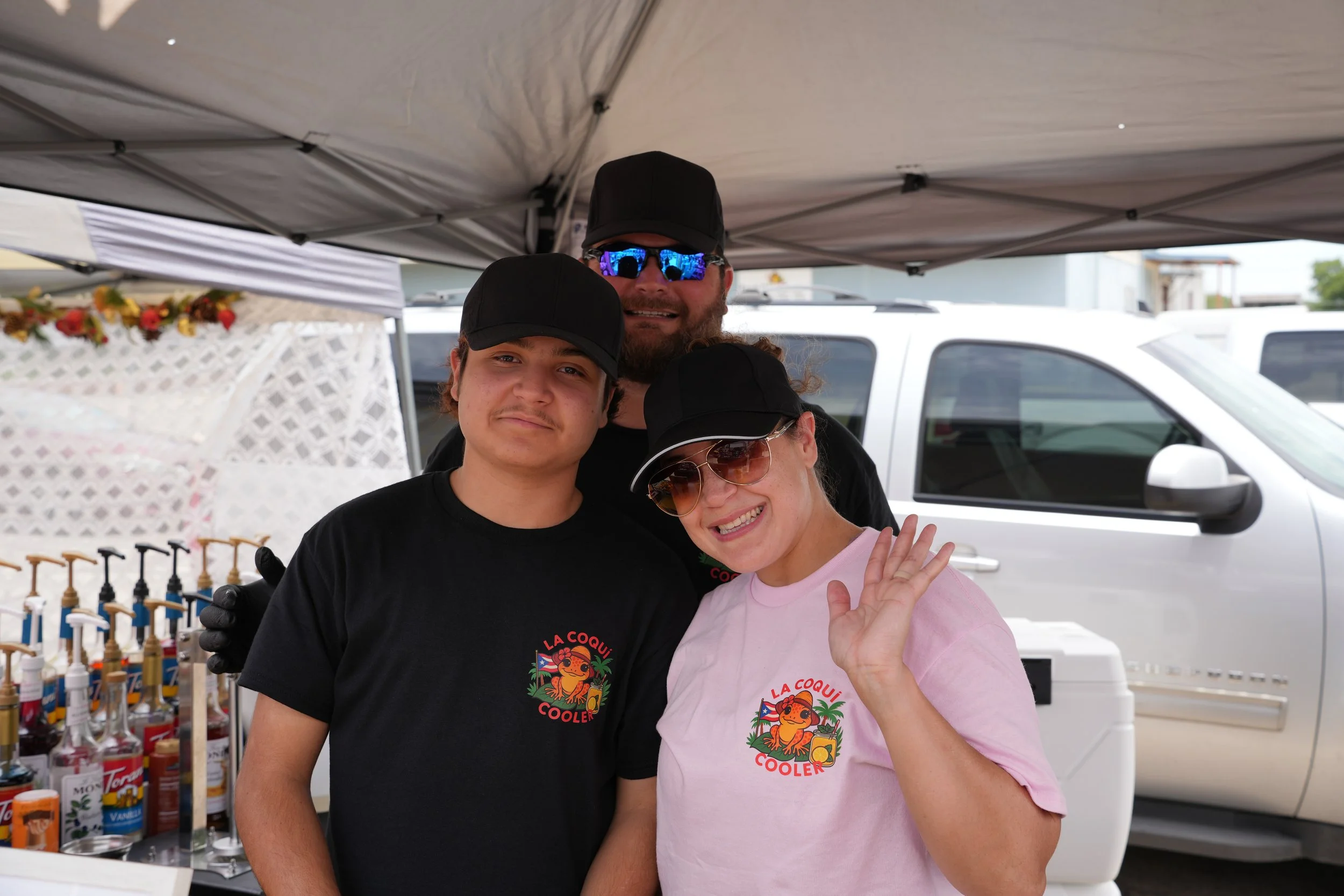 Three people smiling and posing for a photo under a canopy at an outdoor event. Two of them are wearing black caps and shirts with a colorful logo that says 'La Coqui Cooler.' One woman is wearing sunglasses and waving, and a man in the background is wearing sunglasses and a cap.