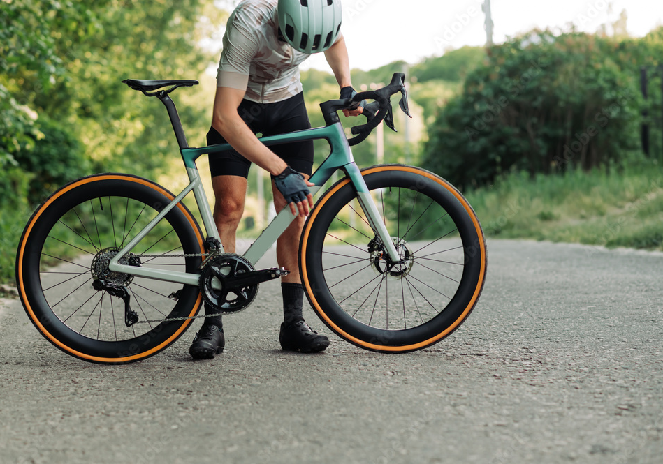 A cyclist wearing a helmet and gloves checking their road bike on a paved trail surrounded by greenery.
