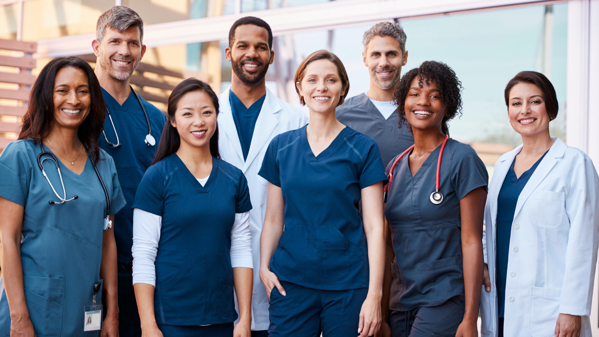 A diverse group of nine healthcare professionals standing together outdoors, smiling and wearing scrubs and lab coats.