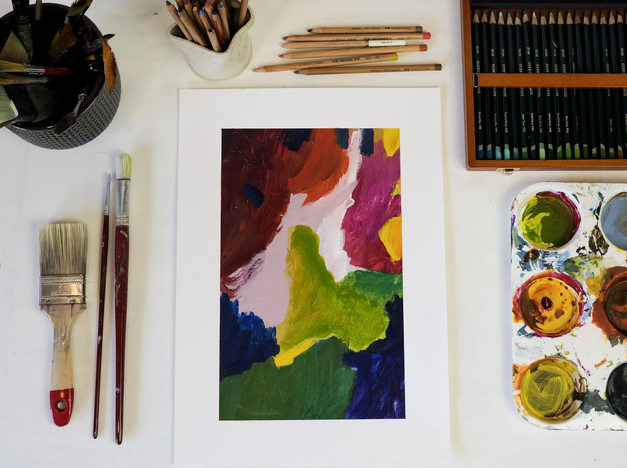 Abstract colorful painting on paper, surrounded by paintbrushes, colored pencils in a container, a palette with various paints, and a box of pencils, on a white table.