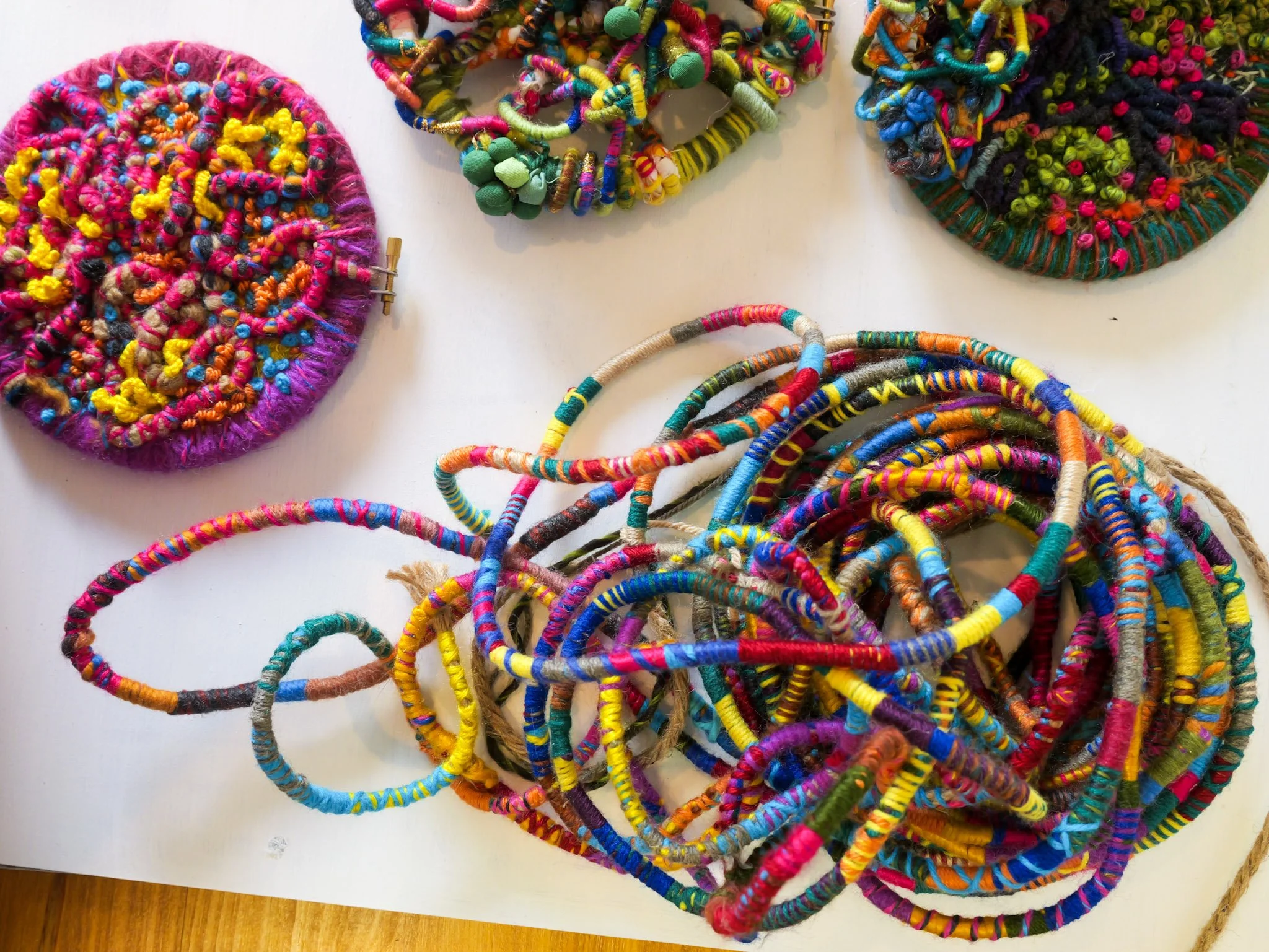 Colorful handmade woven and beaded jewelry, including necklaces and round woven pieces, displayed on a white surface.