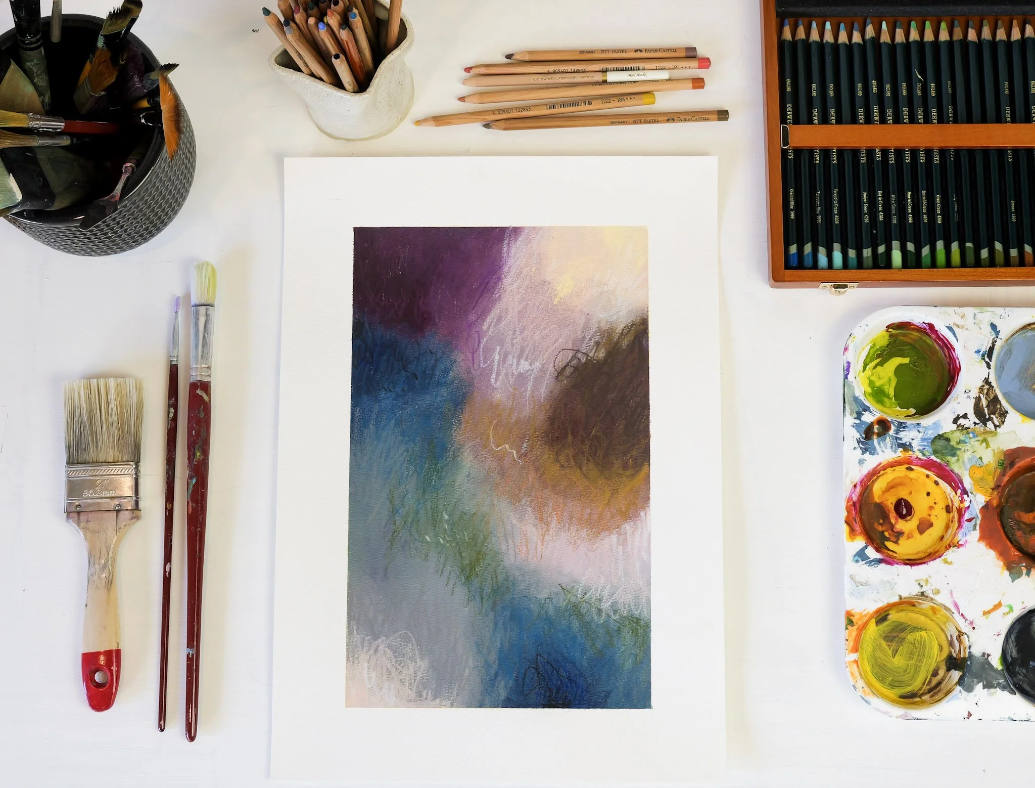 An art workspace with watercolor paints, brushes, colored pencils, and a painting featuring abstract colors and textures.