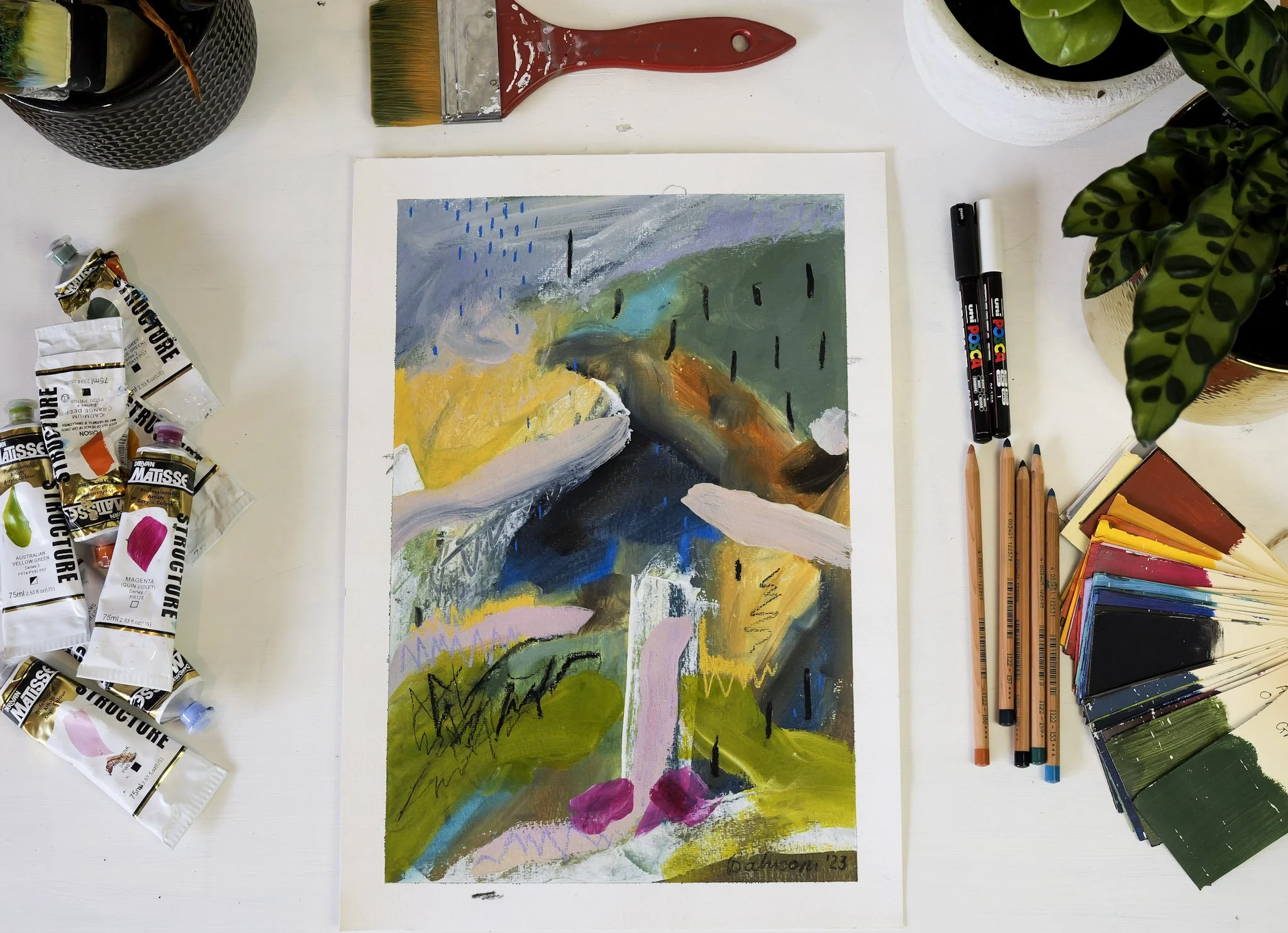 A colorful abstract painting on paper surrounded by various art supplies, including tubes of paint, colored pencils, a color palette, a paintbrush, and two black markers, on a white table.
