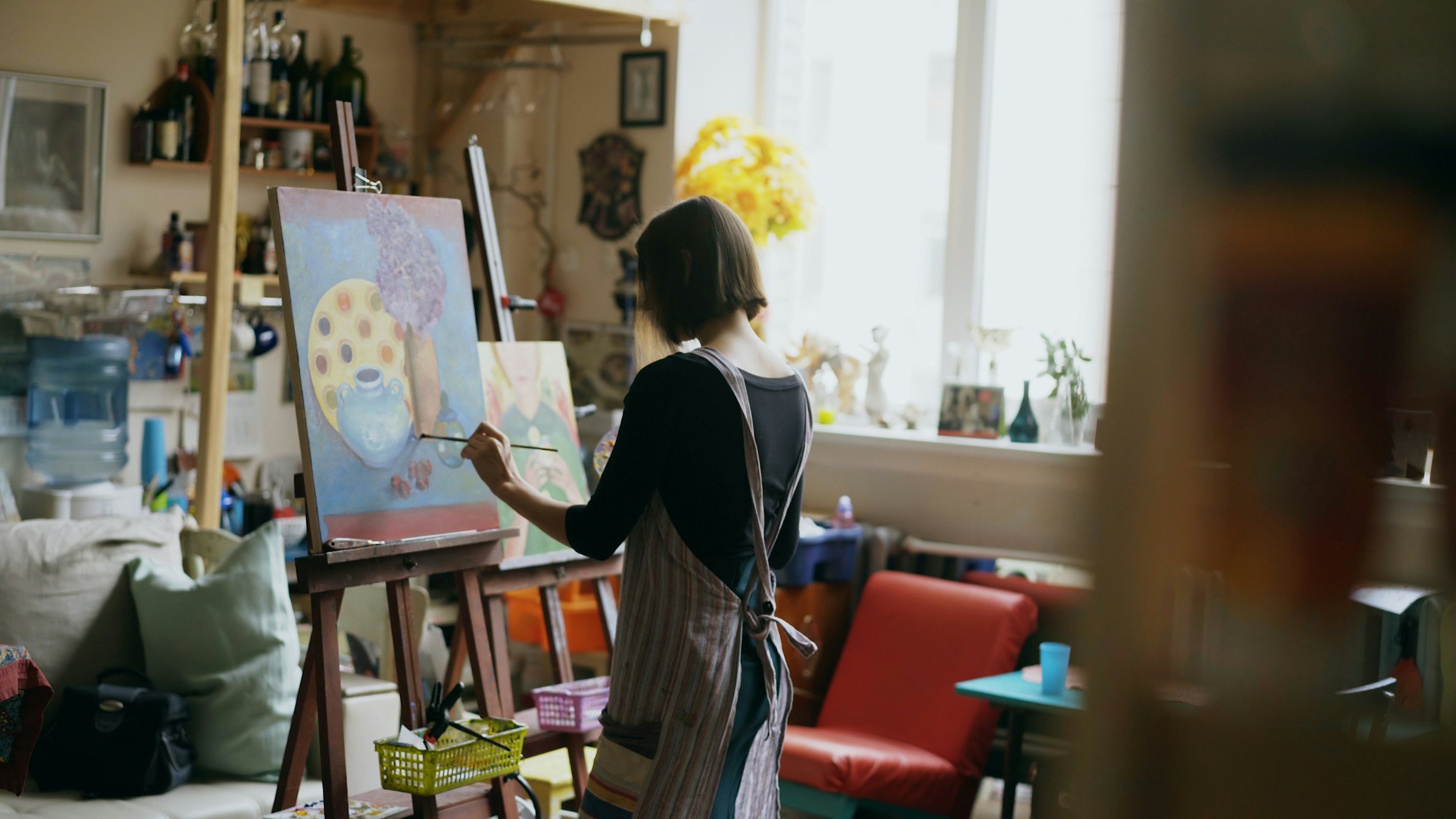 A person wearing a striped apron painting on a canvas in an art studio filled with supplies and artwork.