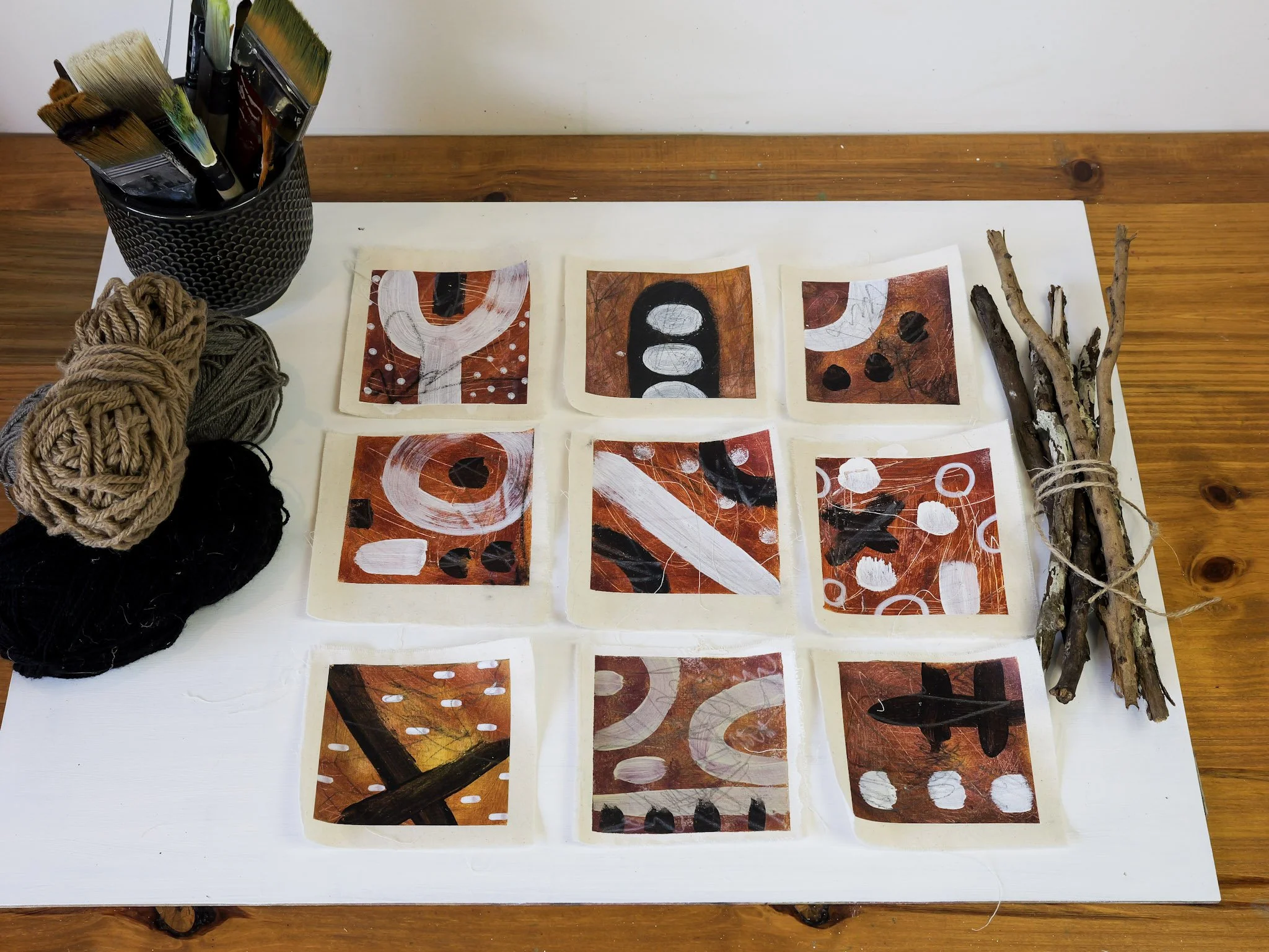 Art supplies and small art pieces on a wooden table, including paintbrushes in a holder, skeins of yarn, and sticks tied with string.