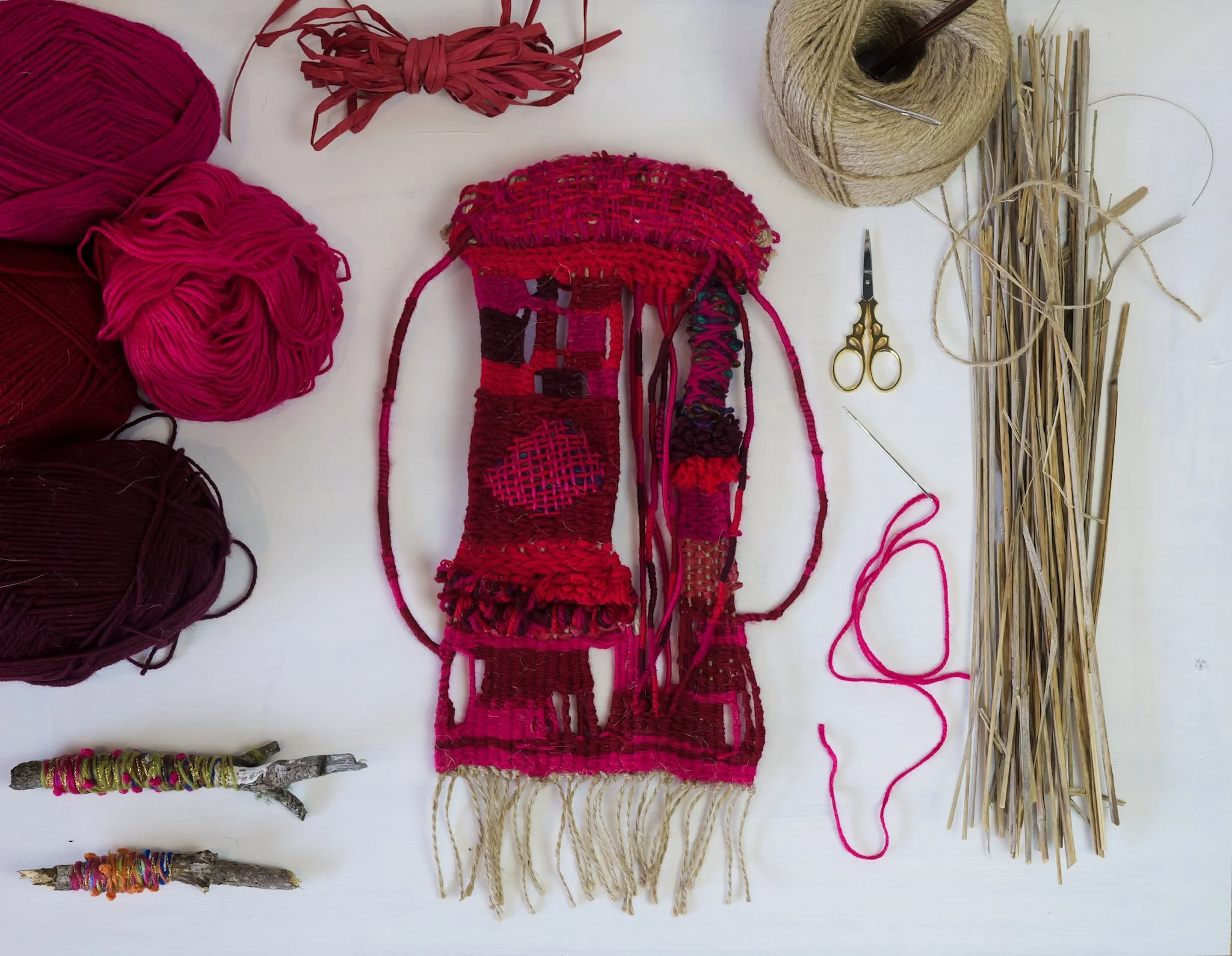 A weaving loom with pink and red yarn, surrounded by balls of yarn in similar shades, a pair of small scissors, spools of thread, and natural fibers, all arranged on a white surface.