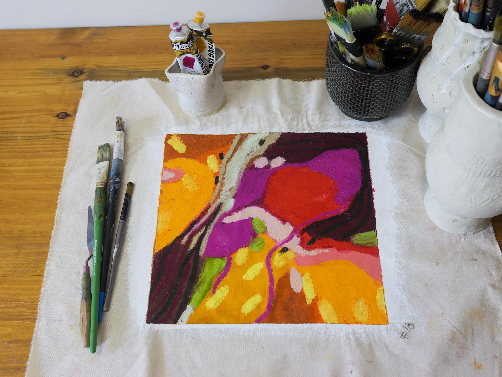 A colorful abstract painting created with acrylics on canvas, placed on a white cloth on a wooden table, surrounded by paintbrushes and containers of paint.