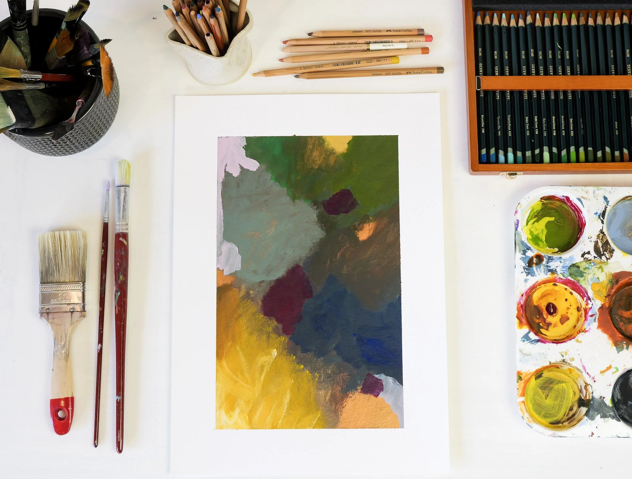 An abstract painting on paper surrounded by art supplies including brushes, colored pencils, watercolors, and a palette with paint.