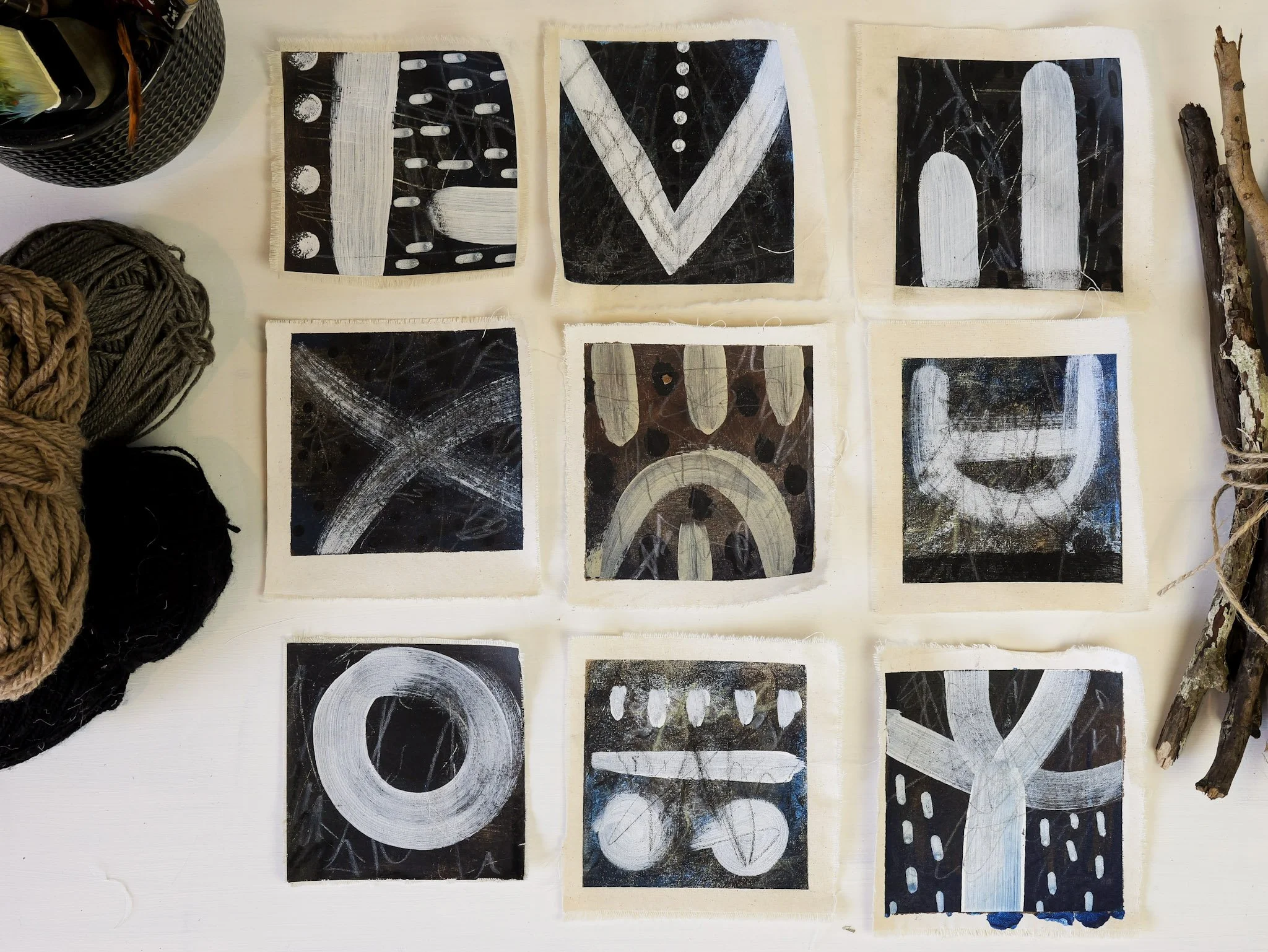 A collection of nine small abstract paintings with black backgrounds and white, beige, and gray brushstrokes, arranged in a 3x3 grid on a white surface, surrounded by yarn balls, sticks, and a black container.