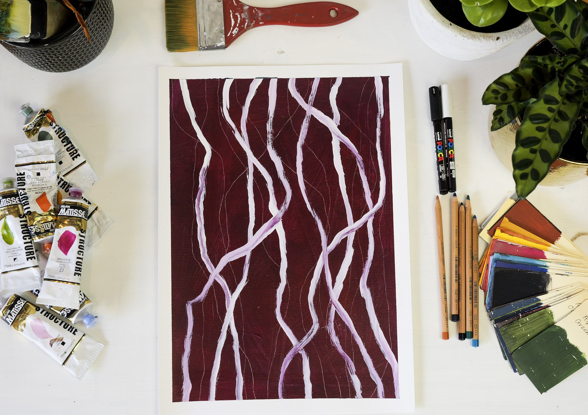 A painting in progress surrounded by art supplies including tubes of paint, paintbrushes, markers, colored paper, and potted plants on a white table.