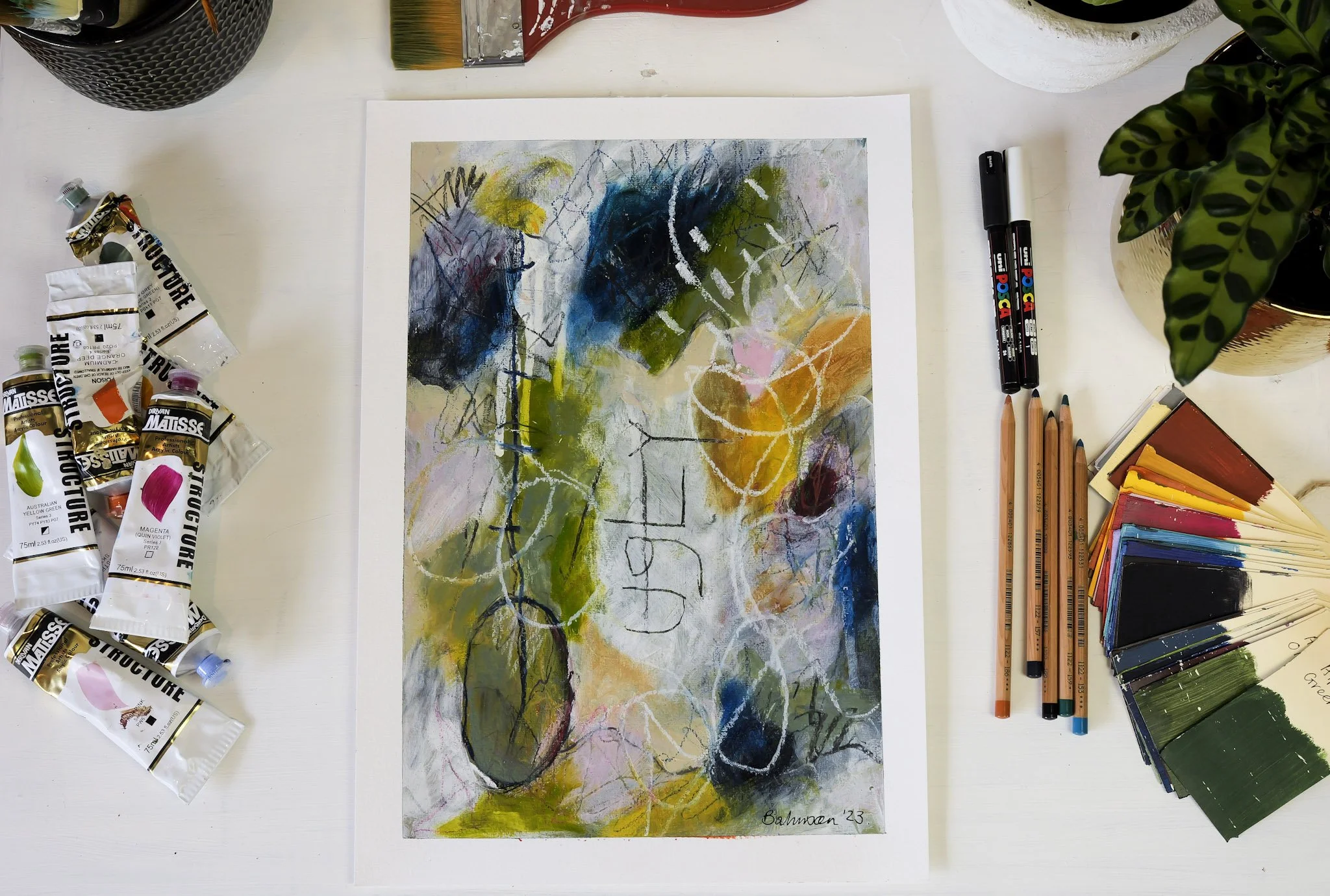 An abstract painting on paper surrounded by watercolor paints, brushes, markers, and a potted plant on a white table.