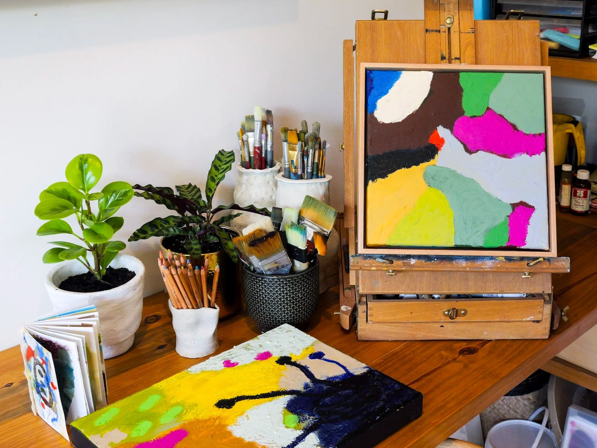 An artist's workspace showing two abstract paintings, various brushes, colored pencils, and plants on a wooden table.