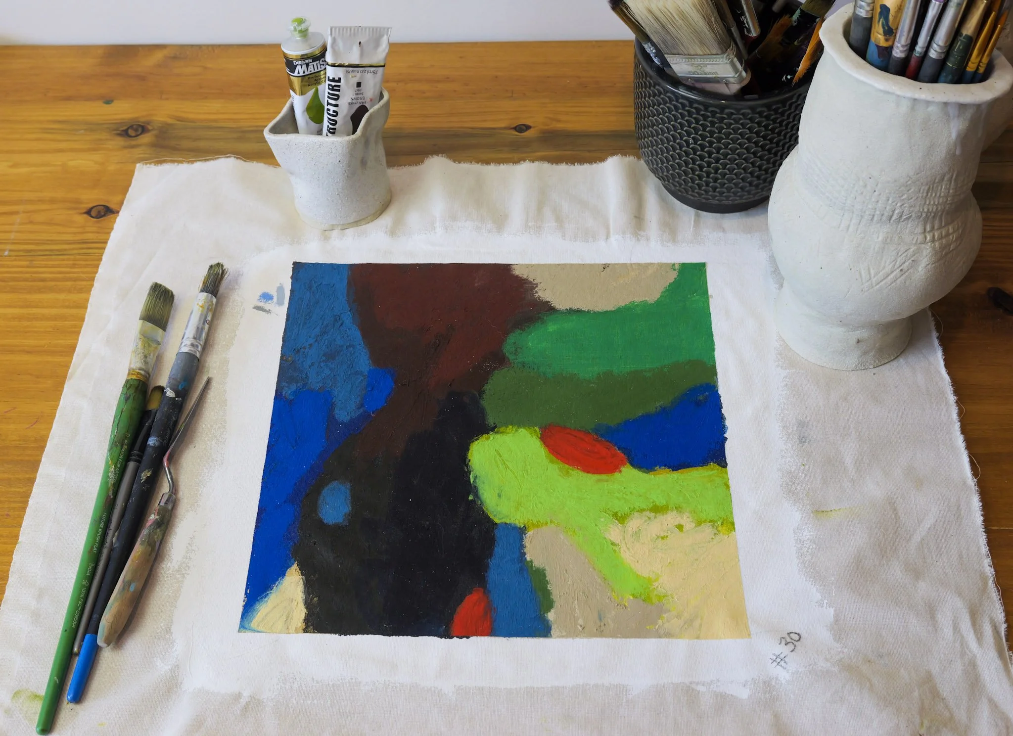 A colorful abstract painting in progress on a cloth-covered table, with paintbrushes, paint tubes, and containers holding art supplies around it.