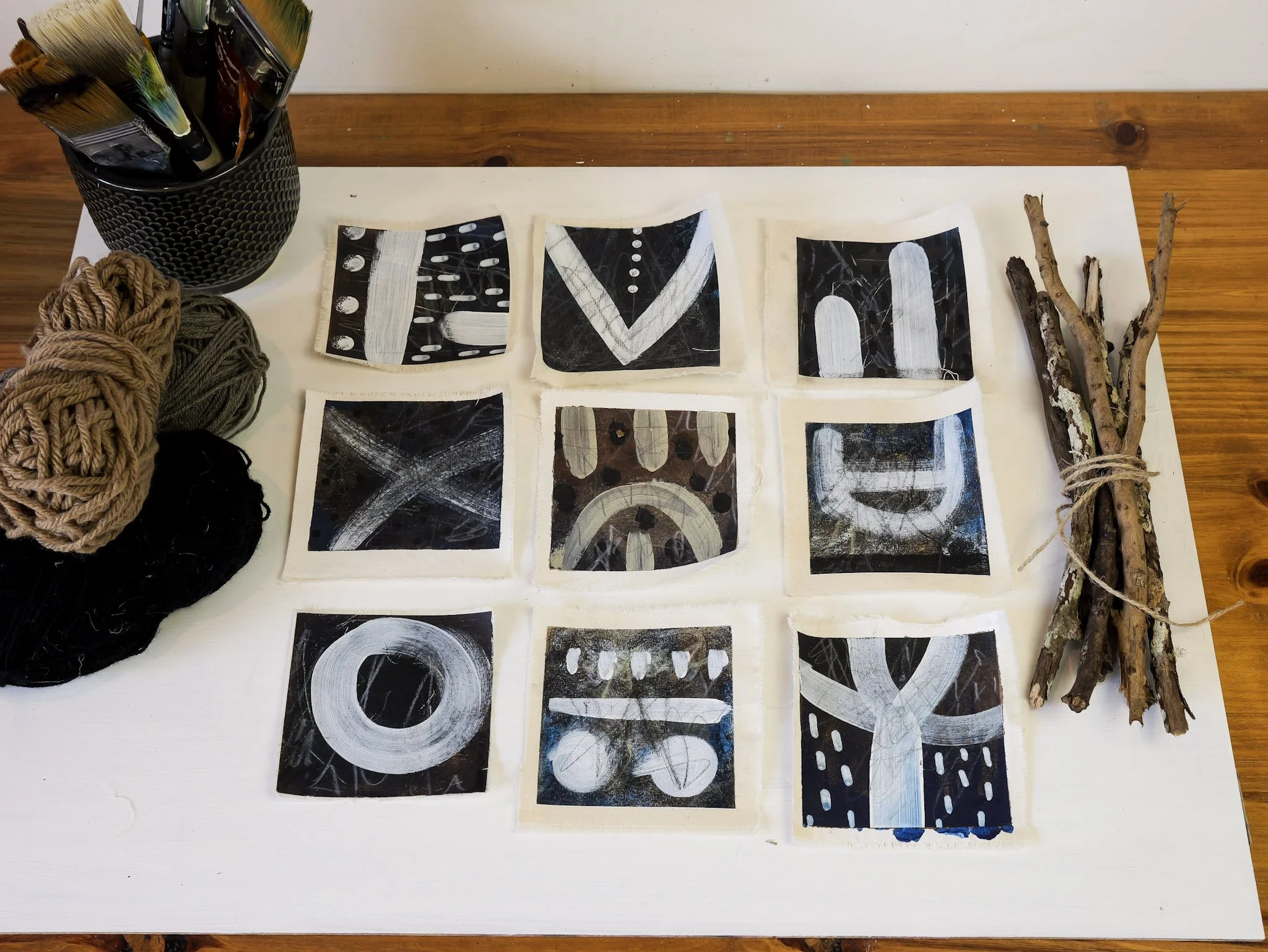 Collection of nine black and white painted tiles arranged on a white surface, with yarn balls and paintbrushes on the left, and a bundle of sticks tied with string on the right.