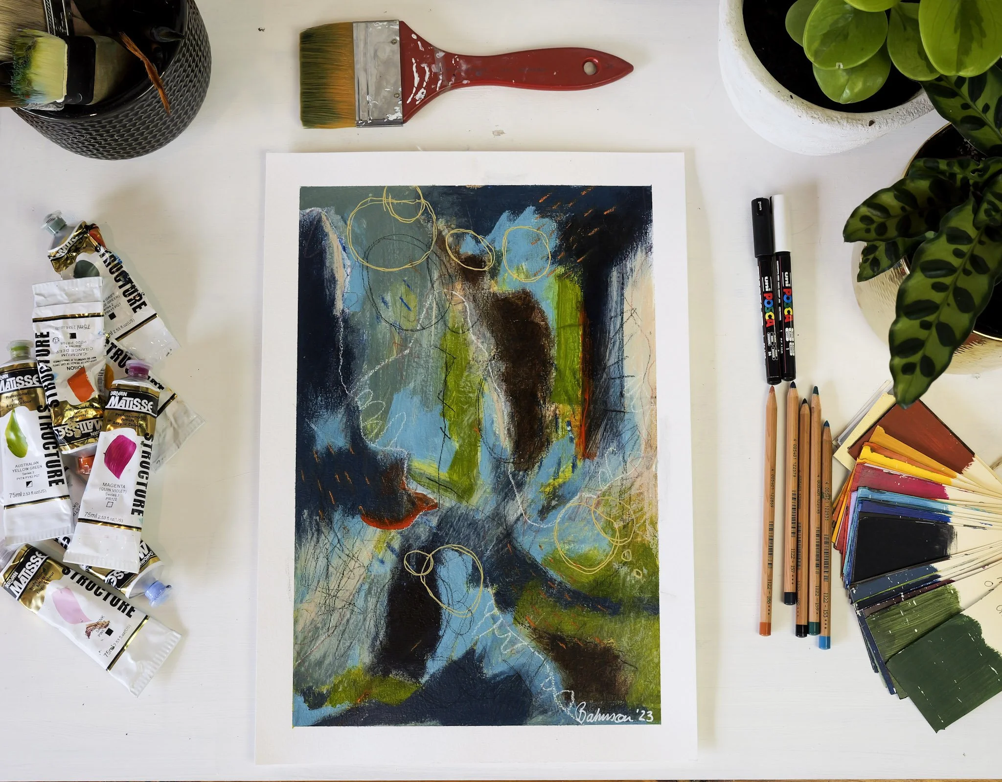 A colorful abstract artwork on paper surrounded by art supplies including paint tubes, colored pencils, markers, a paintbrush, and potted plants on a white table.