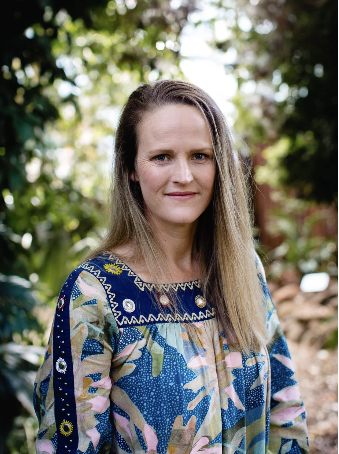Portrait of an artist, wearing a colourful patterned blouse, standing outdoors with greenery and trees in the background.