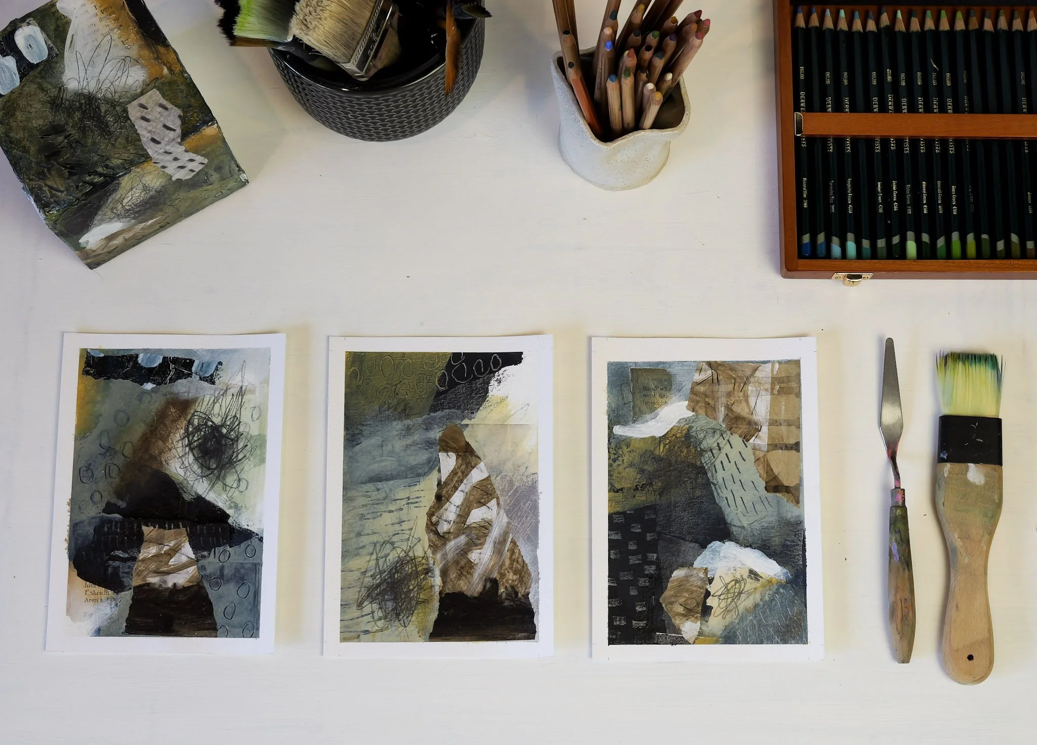 Three collage art pieces laid out on a white table, with art supplies surrounding them, including colored pencils in a wooden box, paintbrushes in a black container, and two palette knives next to them.