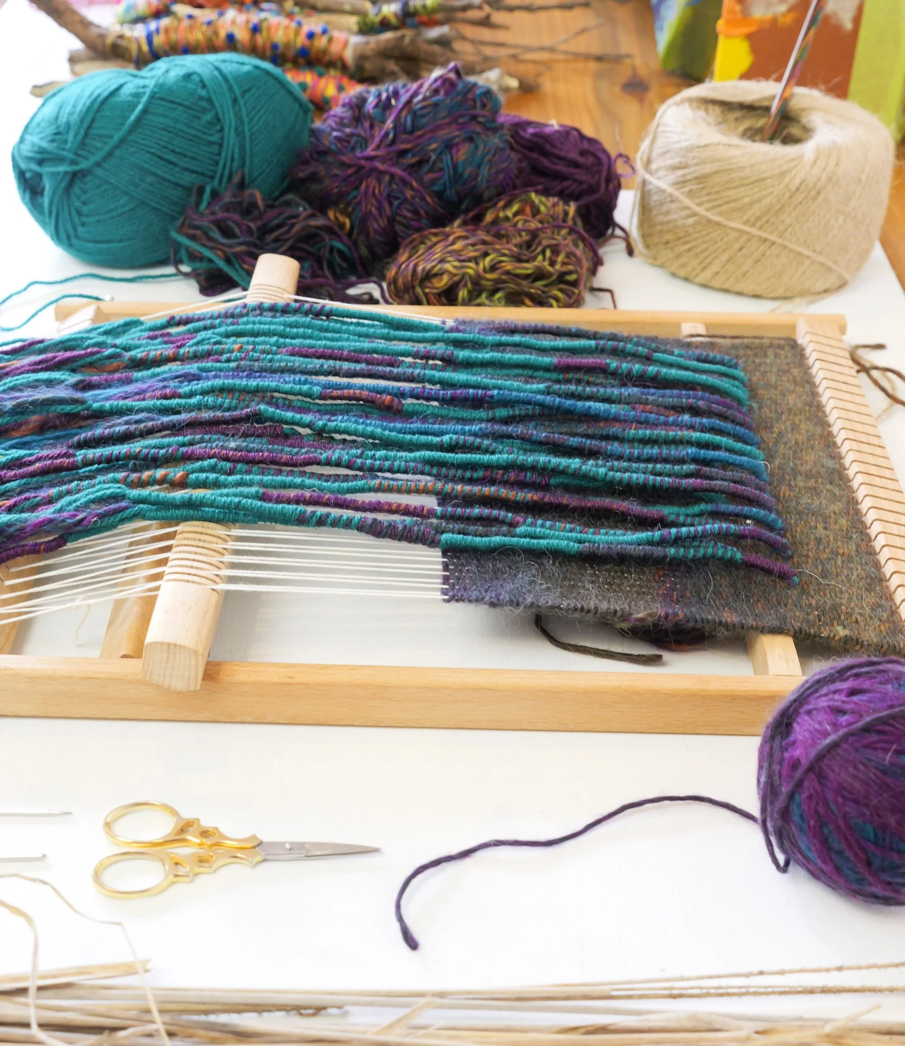 A craft workspace showing colorful yarn, a weaving loom with partially woven fabric, and knitting supplies including scissors, on a white table.