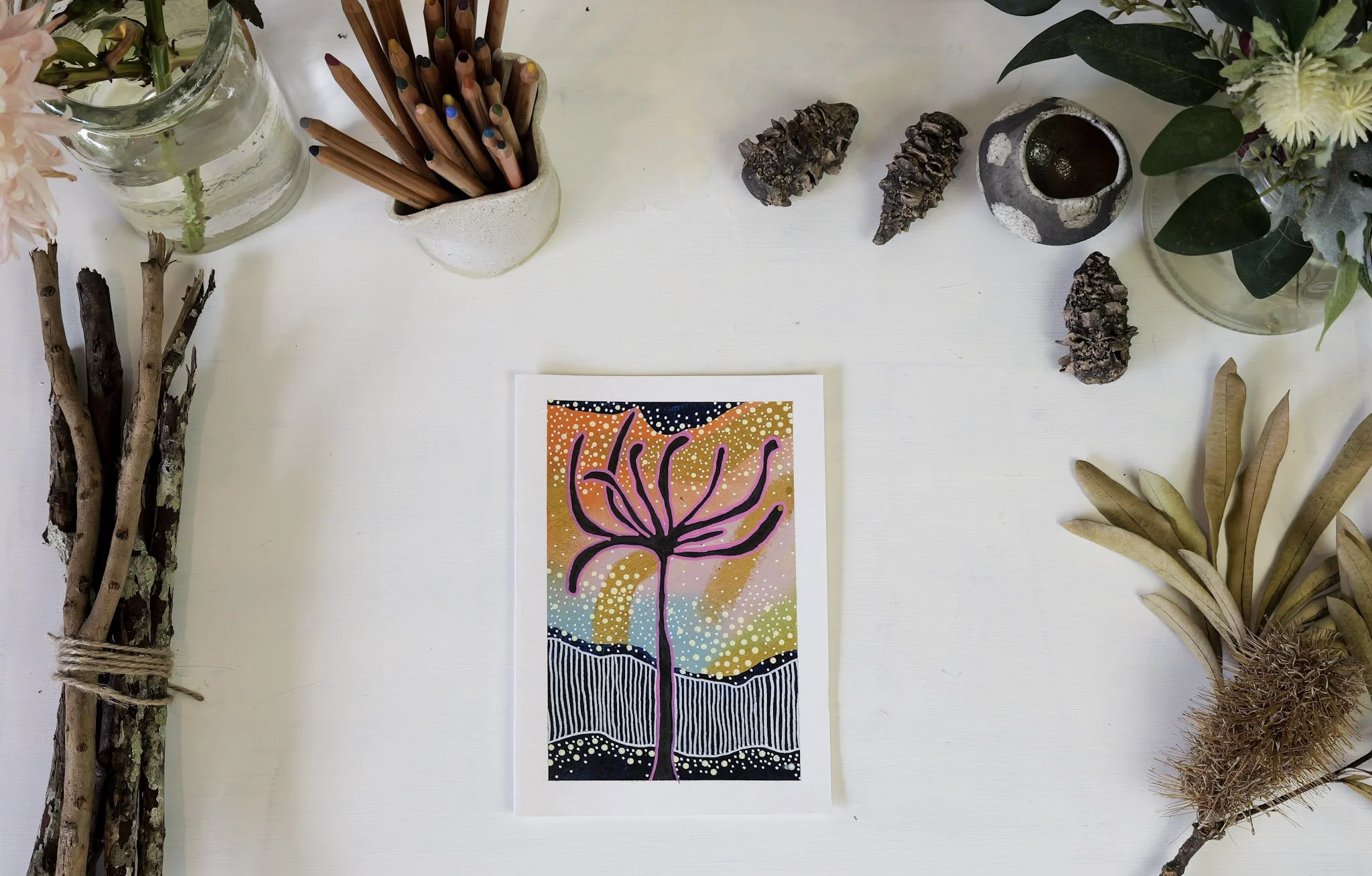 A colorful painting of a tree-like figure with multiple branches on a background of pastel colors and white dots, placed on a white surface. Surrounding items include sticks tied together on the left, colored pencils in a white holder, rocks, a small decorative container, plants, and flowers.