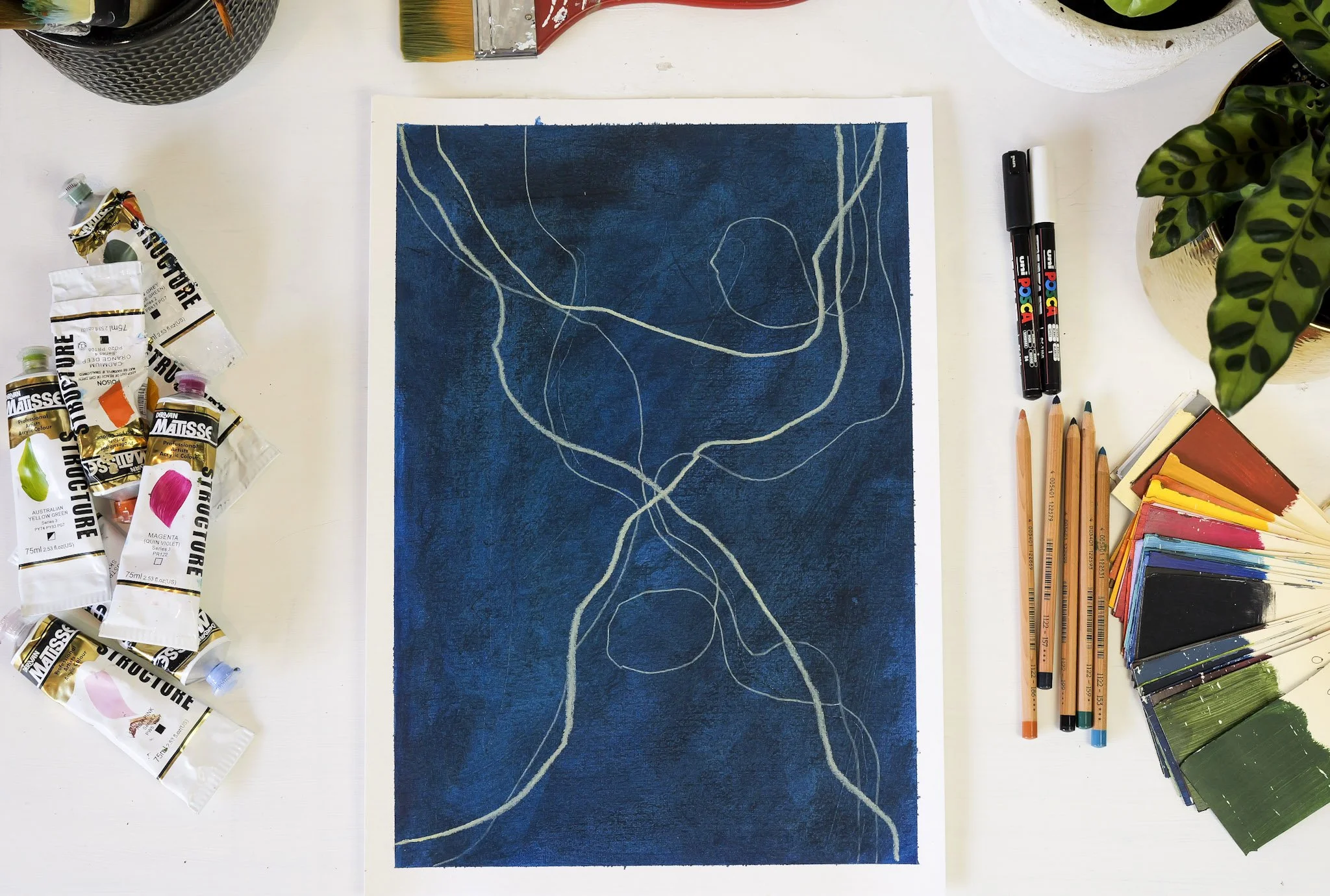 A painting in progress on a white table, featuring a dark blue background with light-colored abstract lines. Surrounding the painting are various art supplies including tubes of watercolor paint, paint swatches, markers, colored pencils, and brushes,