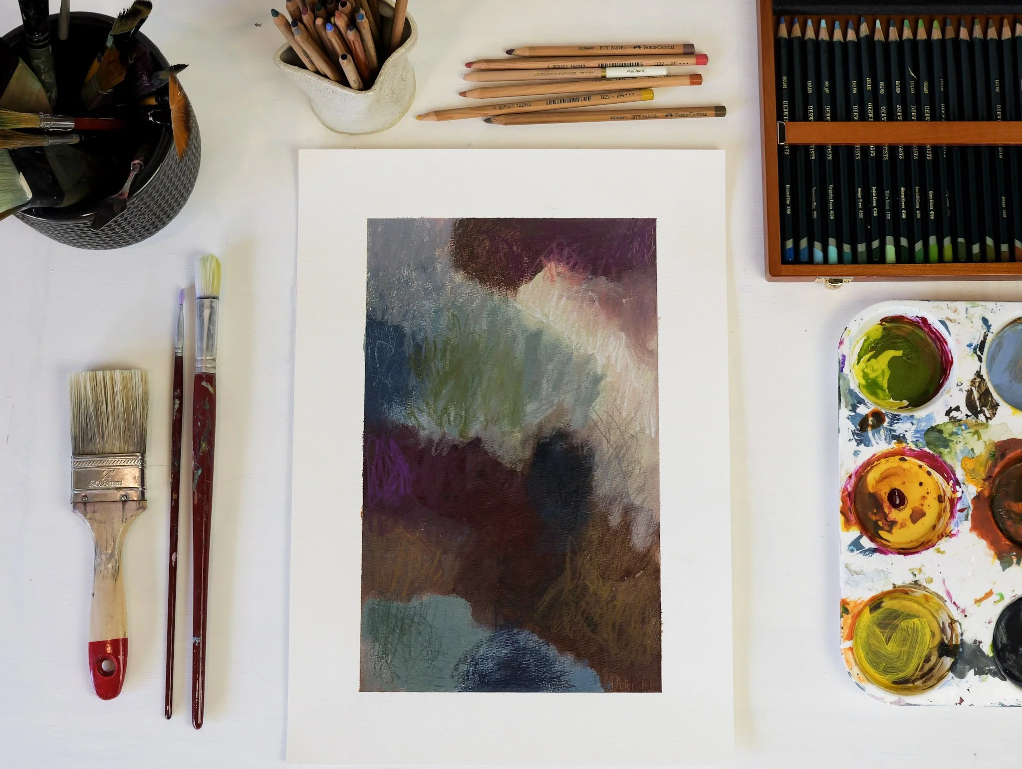 An artist's workspace with a watercolor painting in progress, surrounded by brushes, colored pencils, and paint sets.