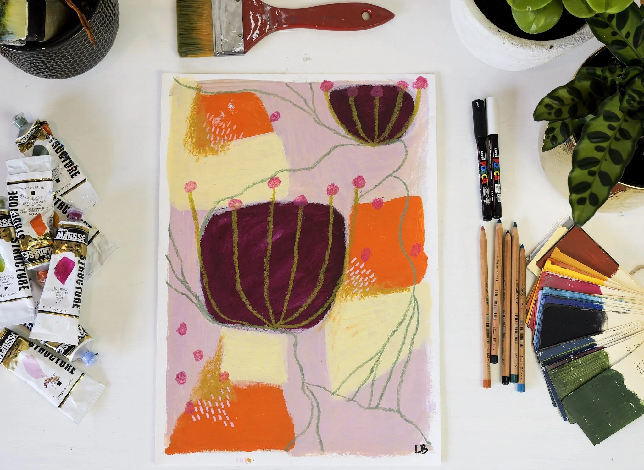 Colorful abstract painting with large purple flowers, orange and yellow shapes, pink dots, and green lines, surrounded by art supplies including paint tubes, brushes, markers, colored pencils, and paint swatches on a white table.