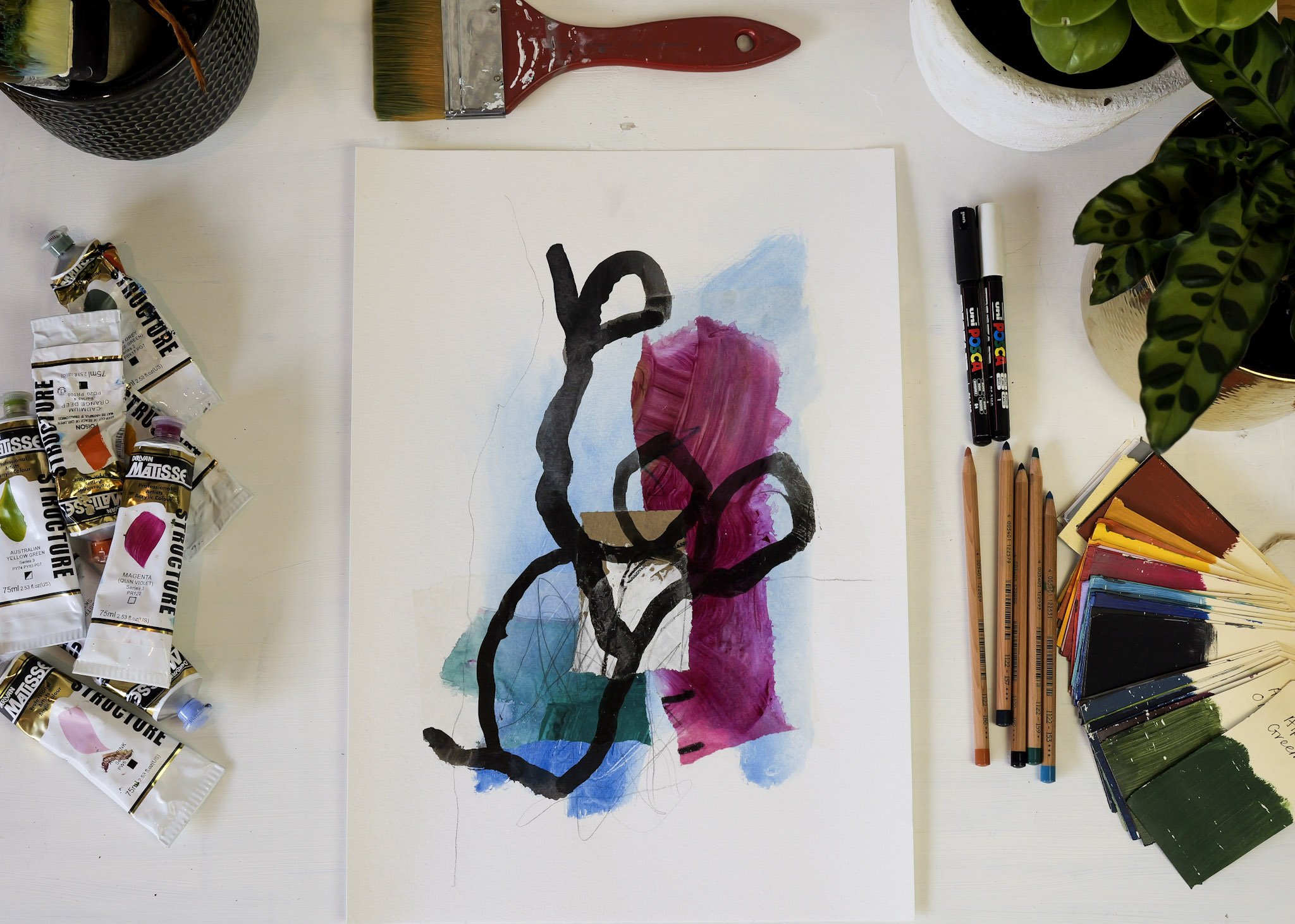 An art workspace with watercolor paints, colored pencils, and a painting in progress on paper, featuring abstract black lines and watercolor splashes of blue, pink, and green.