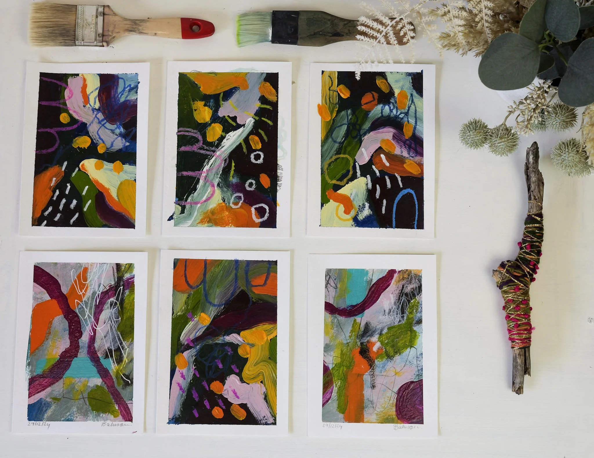 Six colorful abstract paintings arranged in two rows on a white table, with paint brushes and decorative dried plants around them.