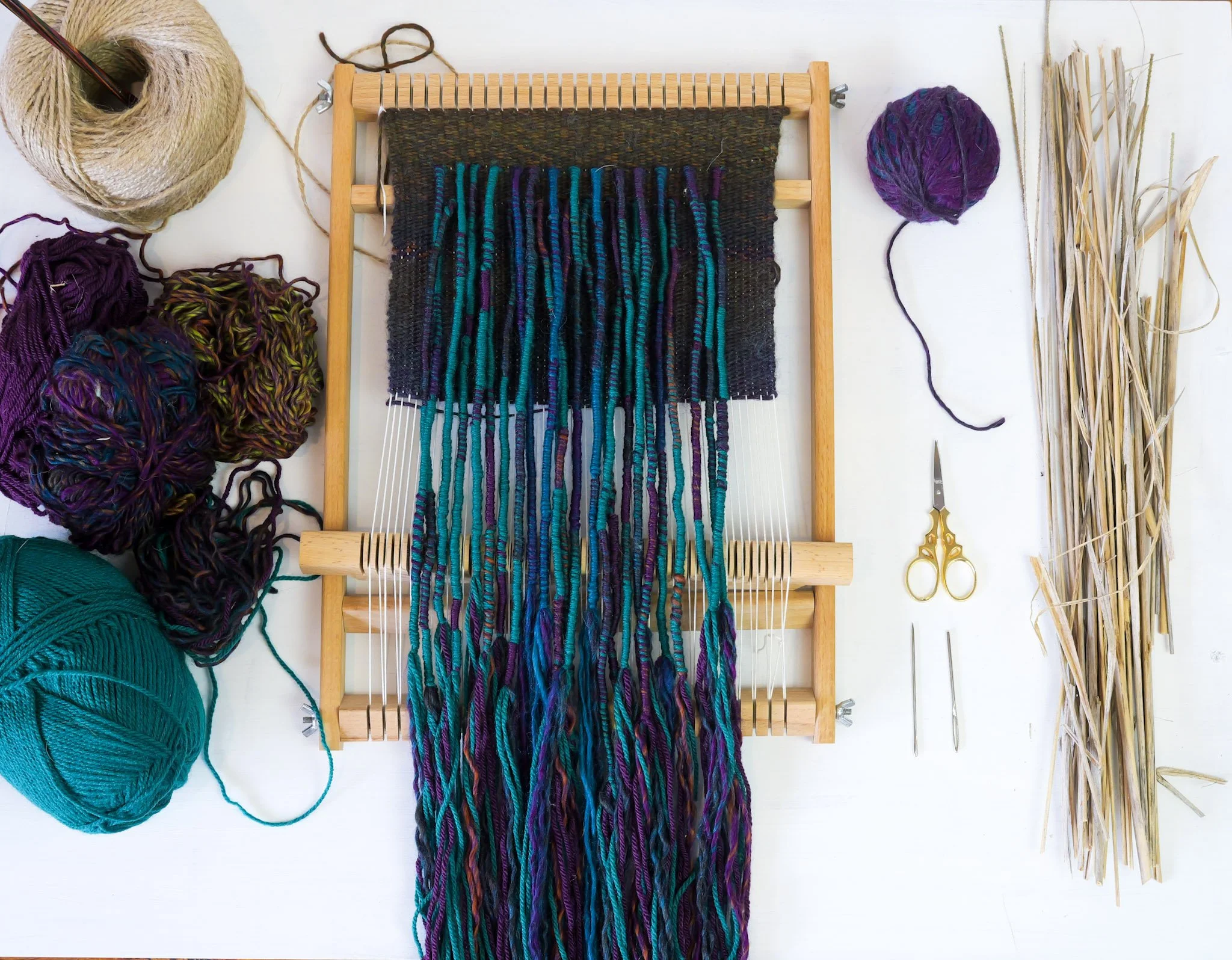 A weaving loom with multicolored yarns and various tools for weaving, including scissors and bundles of dried grass, on a white surface.