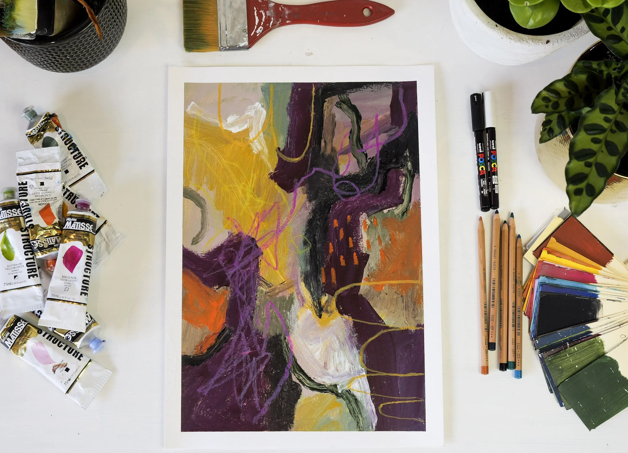 Abstract painting on paper surrounded by art supplies including paint tubes, brushes, colored pencils, markers, color palette, and plants.