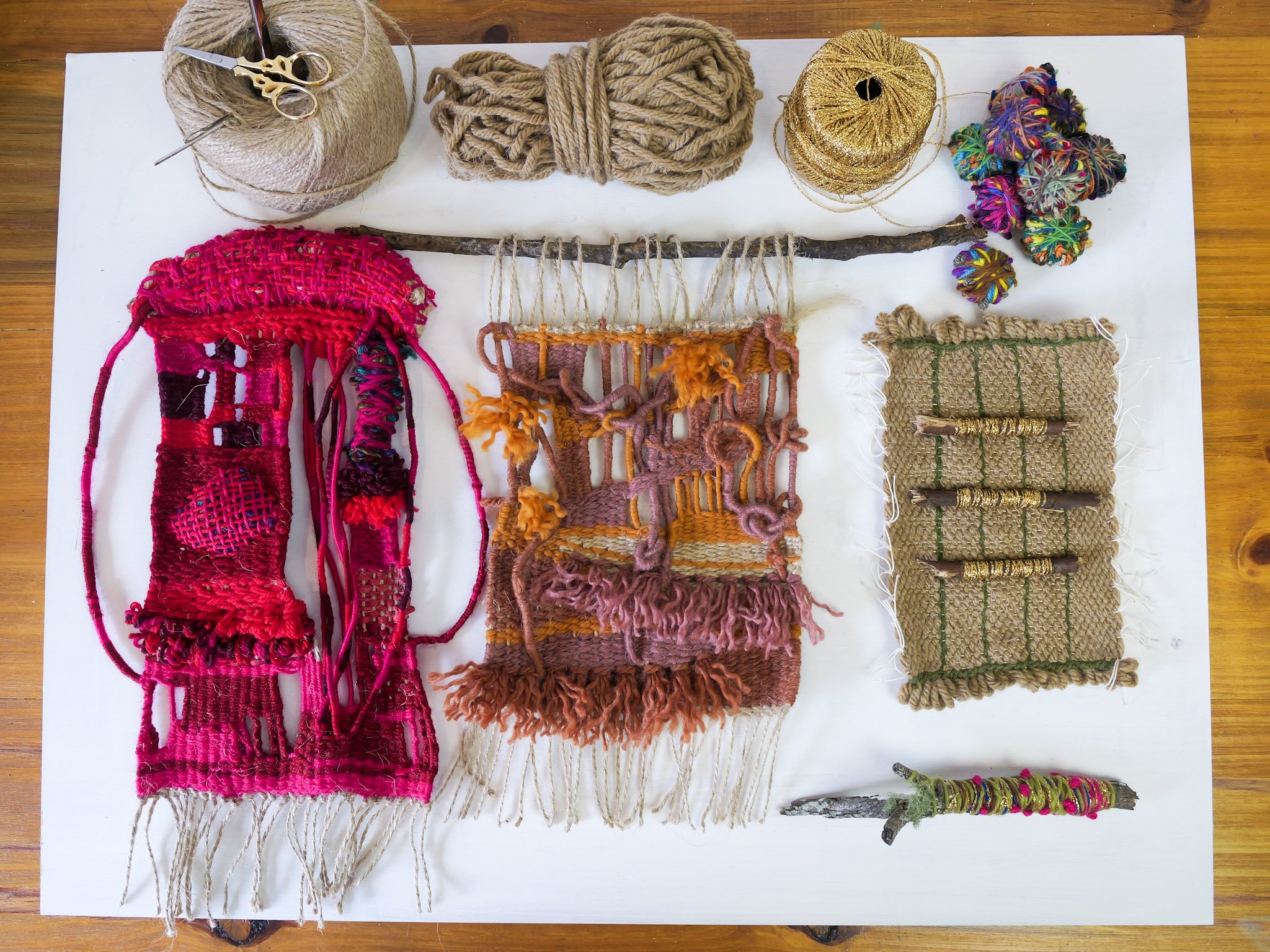 An assortment of colorful woven and textile art pieces on a white surface, including yarn balls, a branch, and various textile samples with fringe and knitting details.