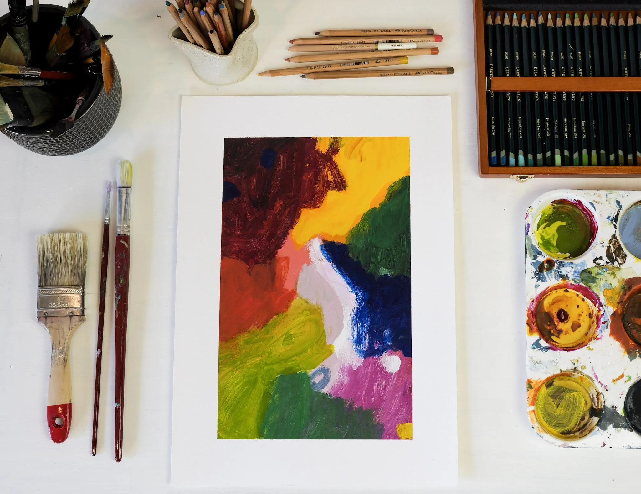 Colorful abstract painting surrounded by paintbrushes, colored pencils, and paint palettes on a white table.