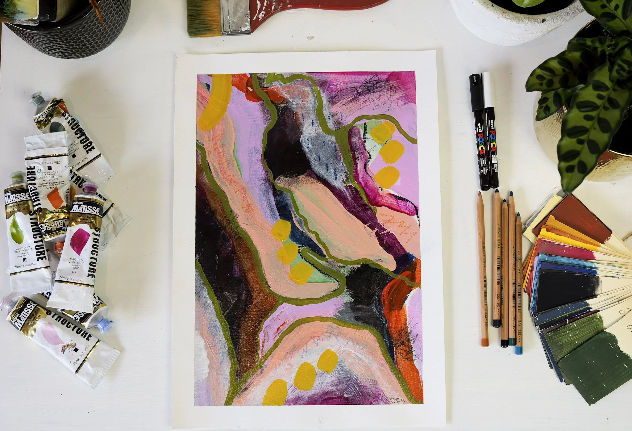 An abstract painting with pink, purple, black, orange, green, and yellow colors on paper, surrounded by art supplies including watercolor tubes, colored pencils, markers, and paint swatches on a white table.